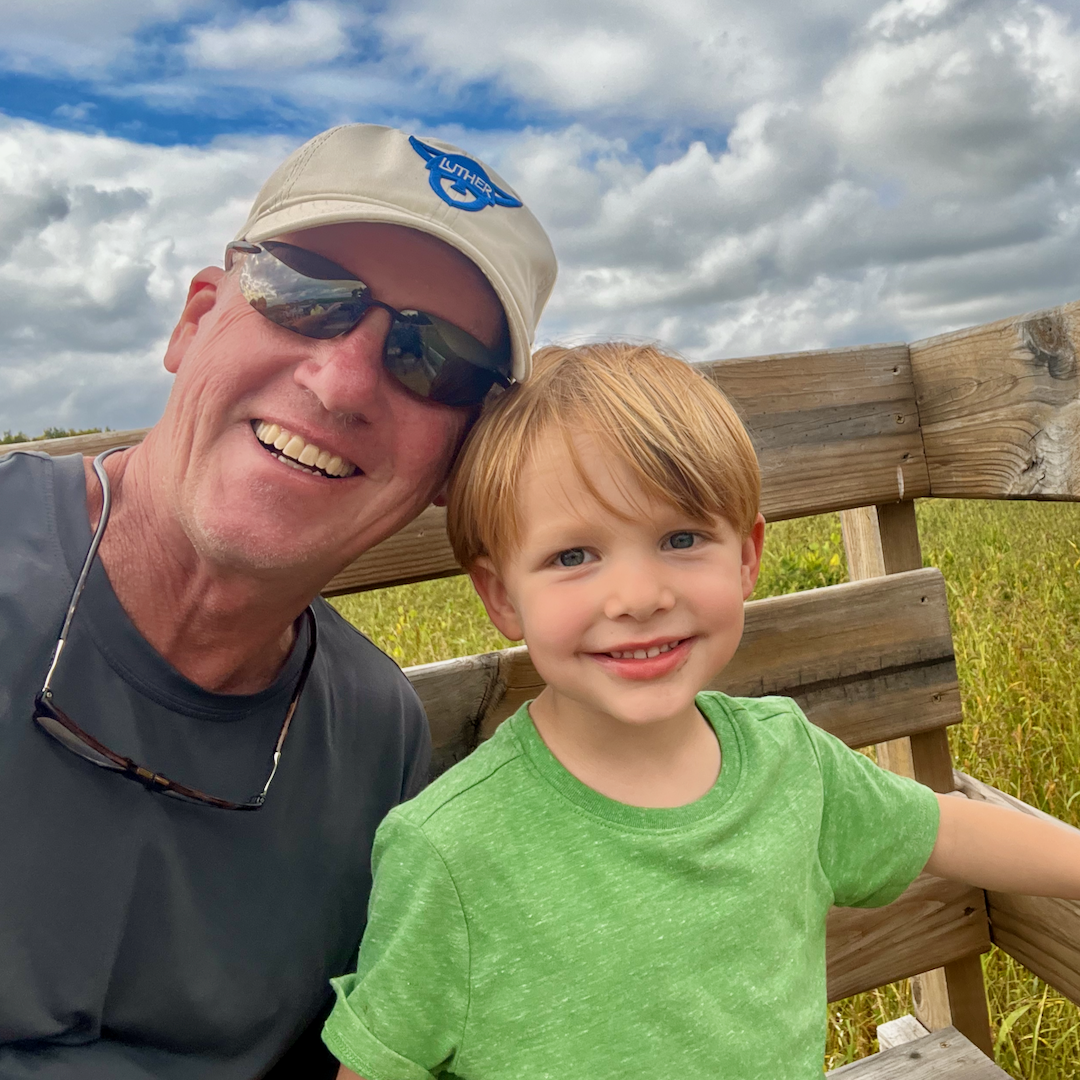 A man wearing sunglasses and a cap poses with a smiling child in a green shirt, sitting on a wooden bench with a cloudy sky in the background.