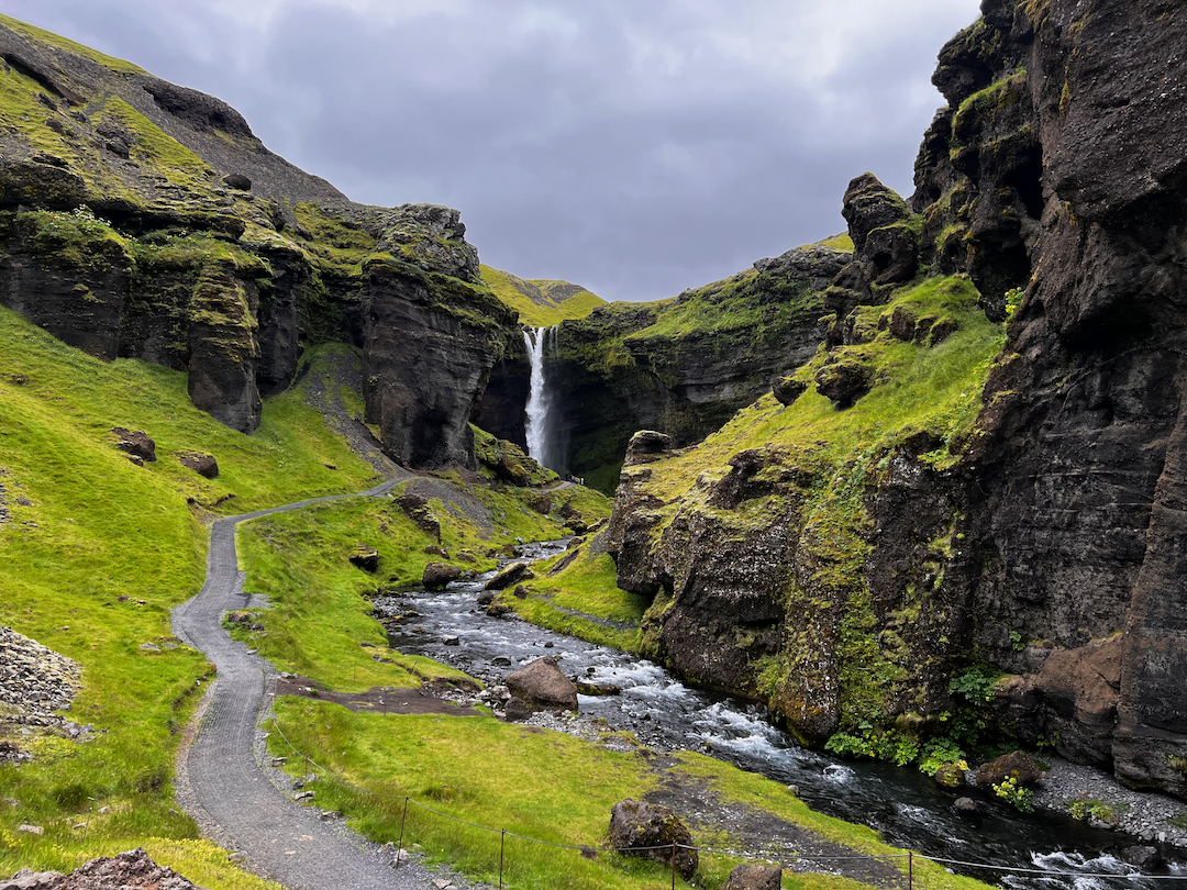 A waterfall flows between lush green cliffs with a winding path and stream in the foreground under a cloudy sky.