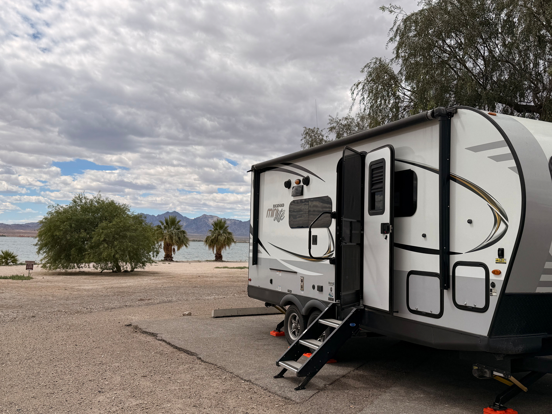 Auto-generated description: A travel trailer is parked near a scenic lakeside area with trees and mountains under a partly cloudy sky.