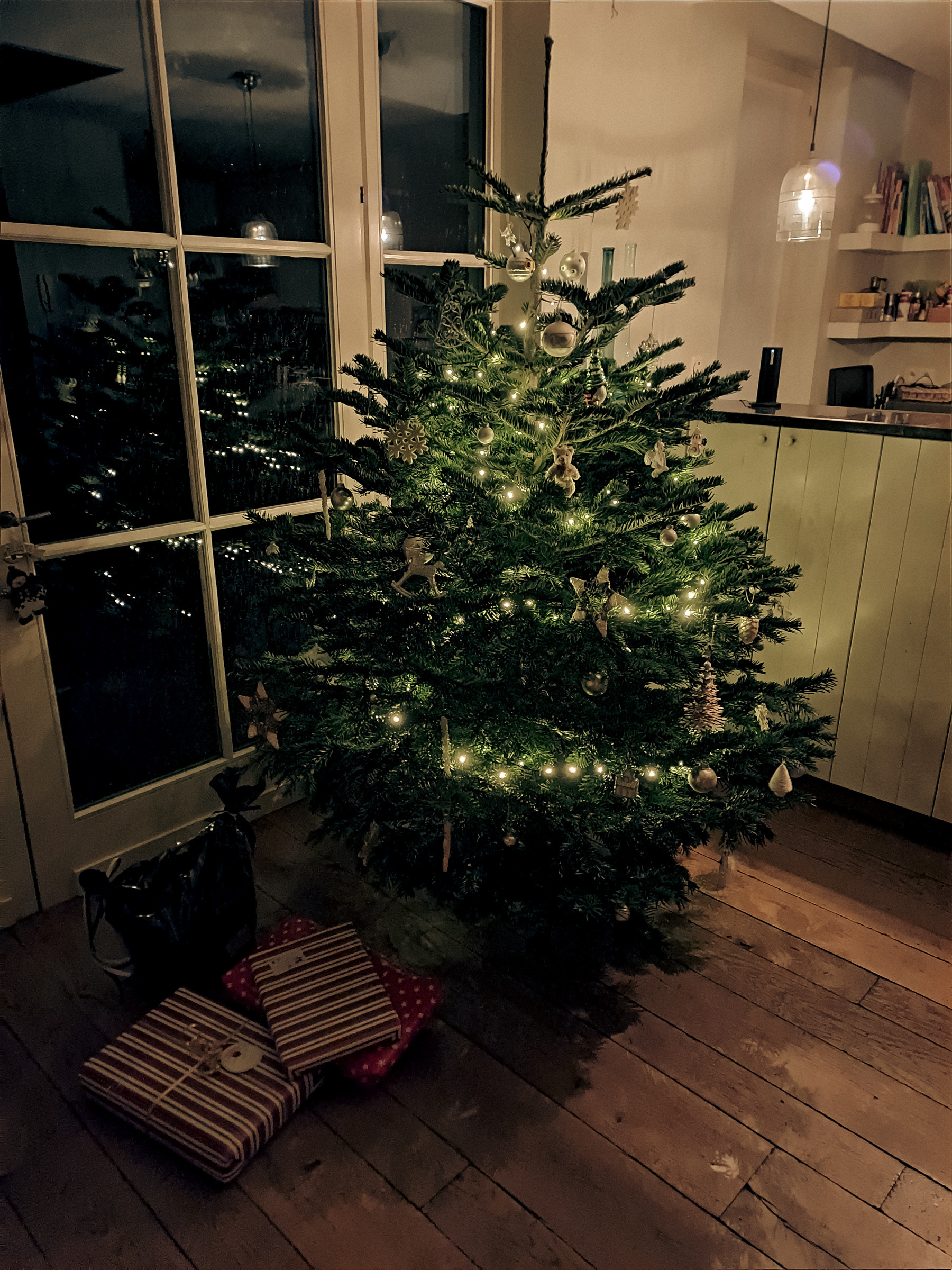 A decorated Christmas tree in the corner of a room, before a lot of glass, some gifst are laying under the tree
