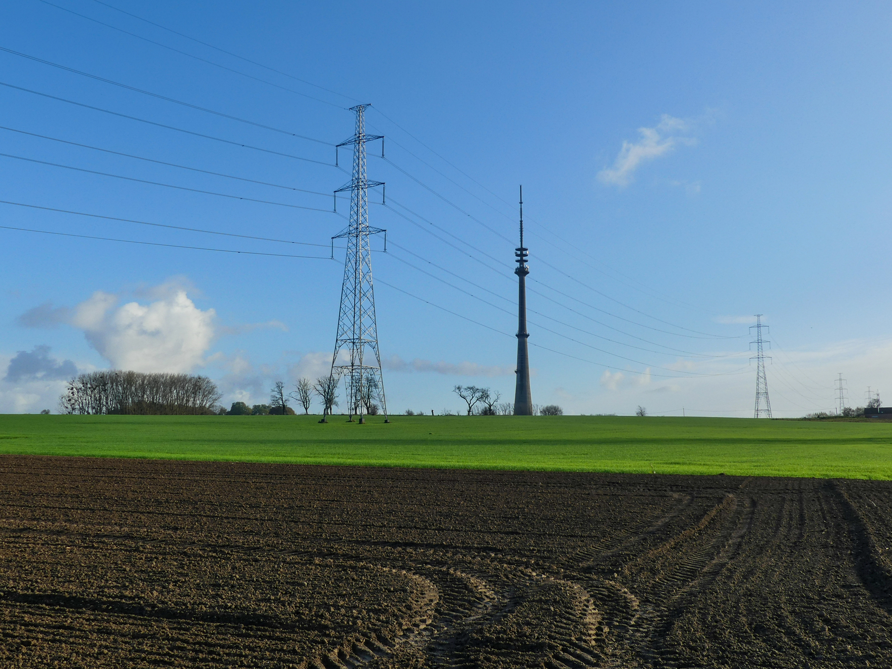 De zendmast van Sint-Pieters-Leeuw omringt door twee elektriciteitsmasten in een groen veld, met omgewoelde aarde op de voorgrond