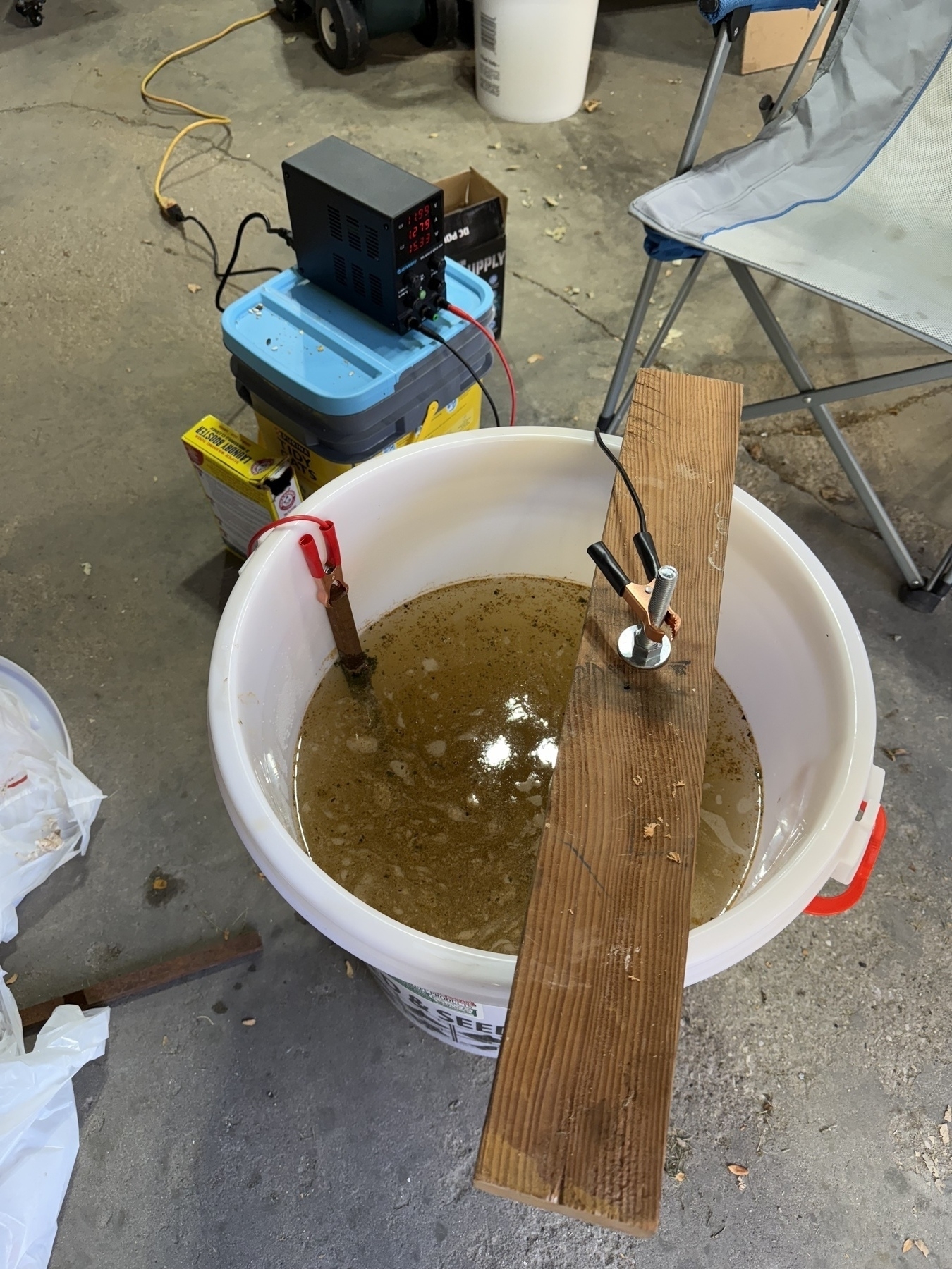 A large white plastic bucket mostly full of dirty water, with a power supply wired up to a piece of steel going into the bucket and also to a cast iron skillet hanging from above the bucket. 