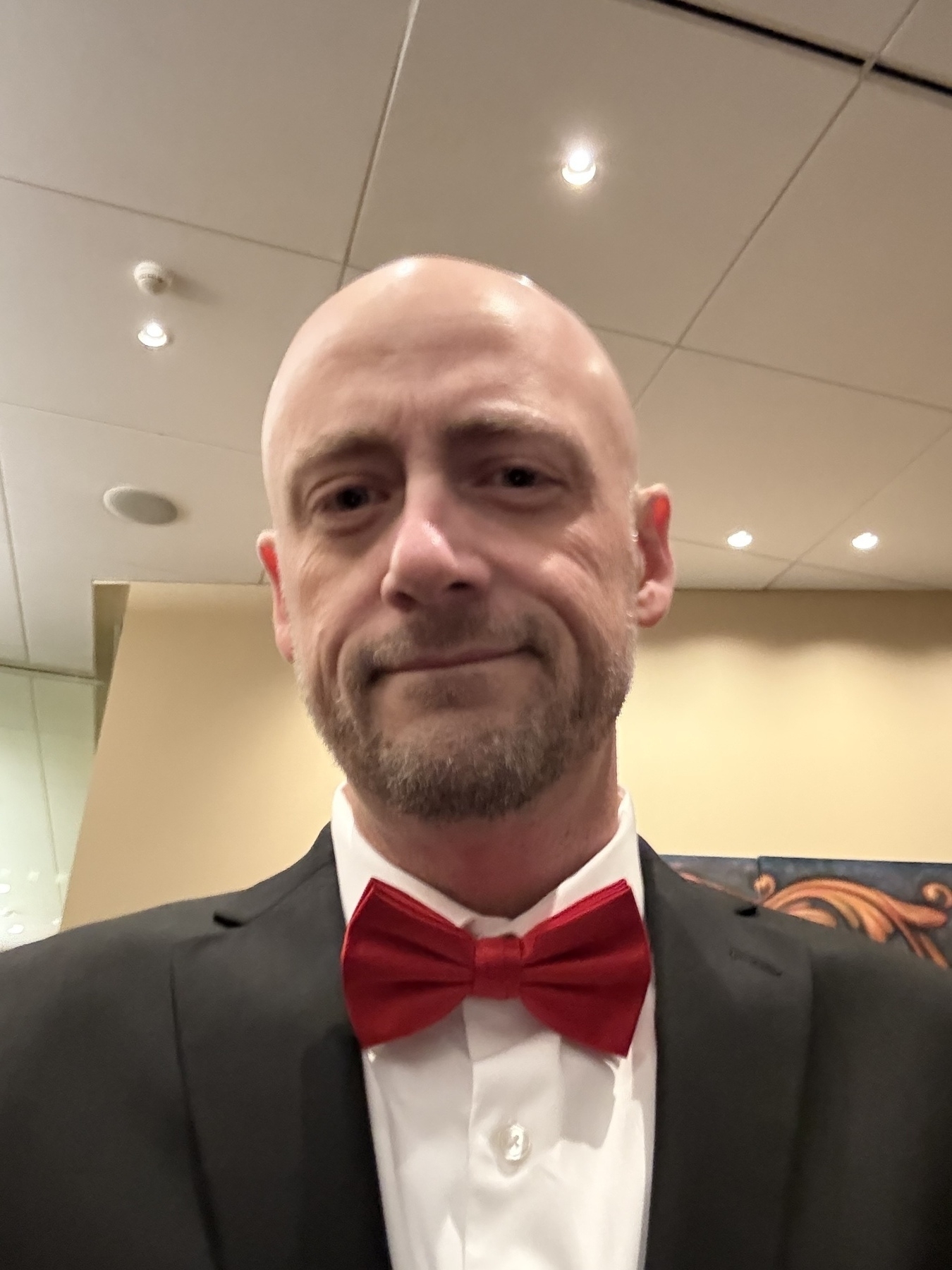 A selfie of a bald, bearded man in a black suit with a red bow tie