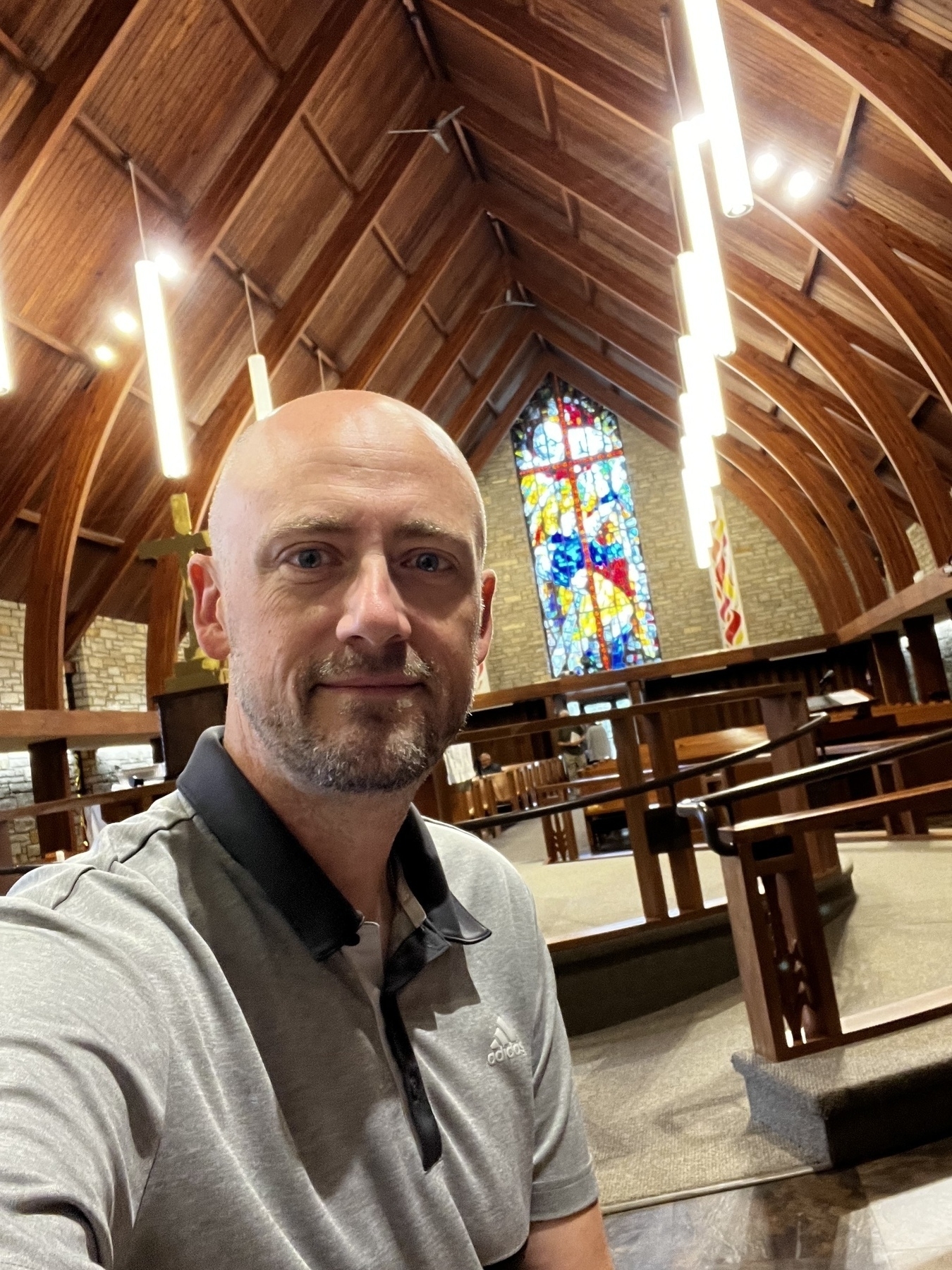 A bald, bearded man in a grey polo shirt with a vaulted church ceiling and stained glass window in the background