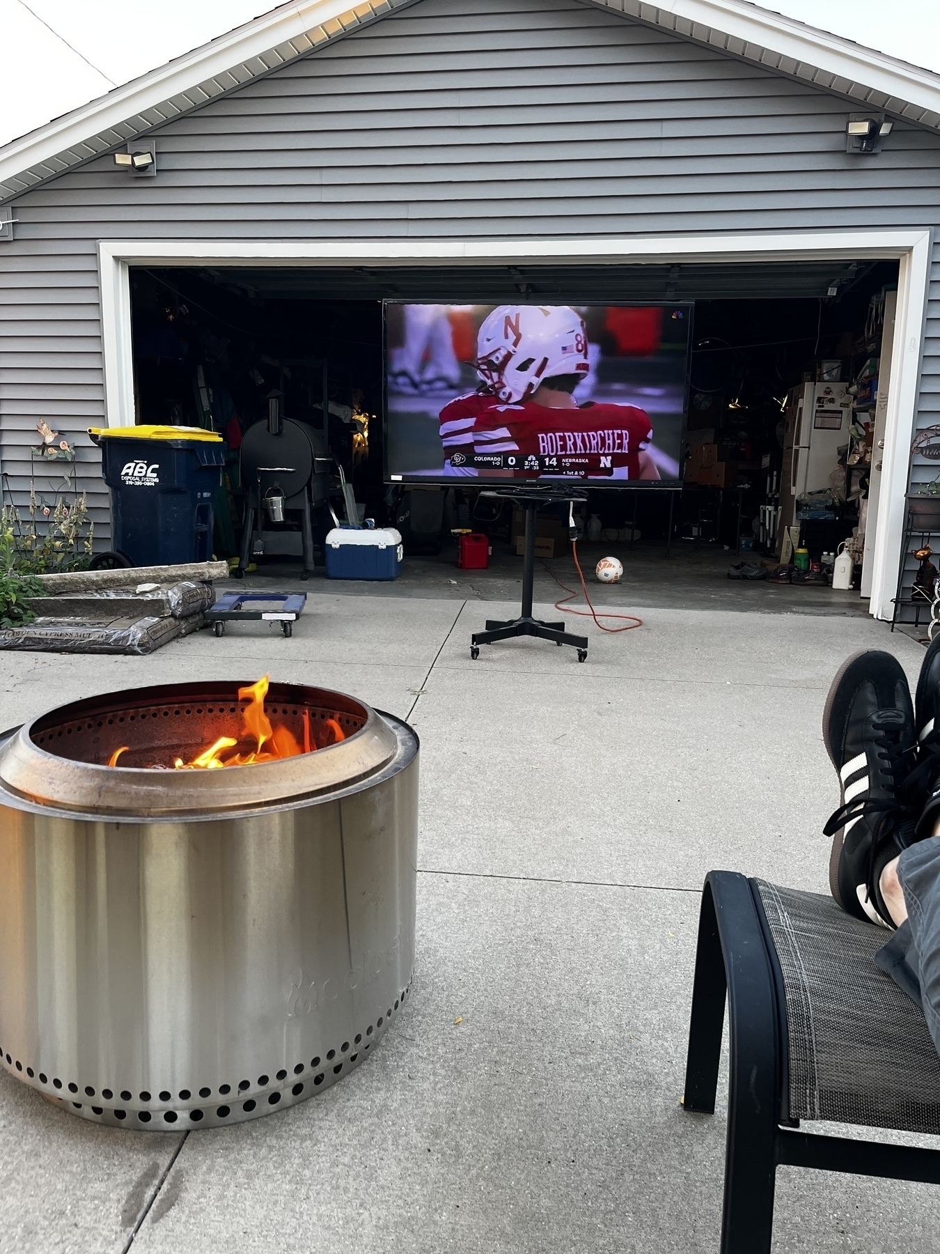 A grey garage with the door open, a silver solo stove with a fire crackling in it, and a big TV in the garage door opening with Nebraska football on it