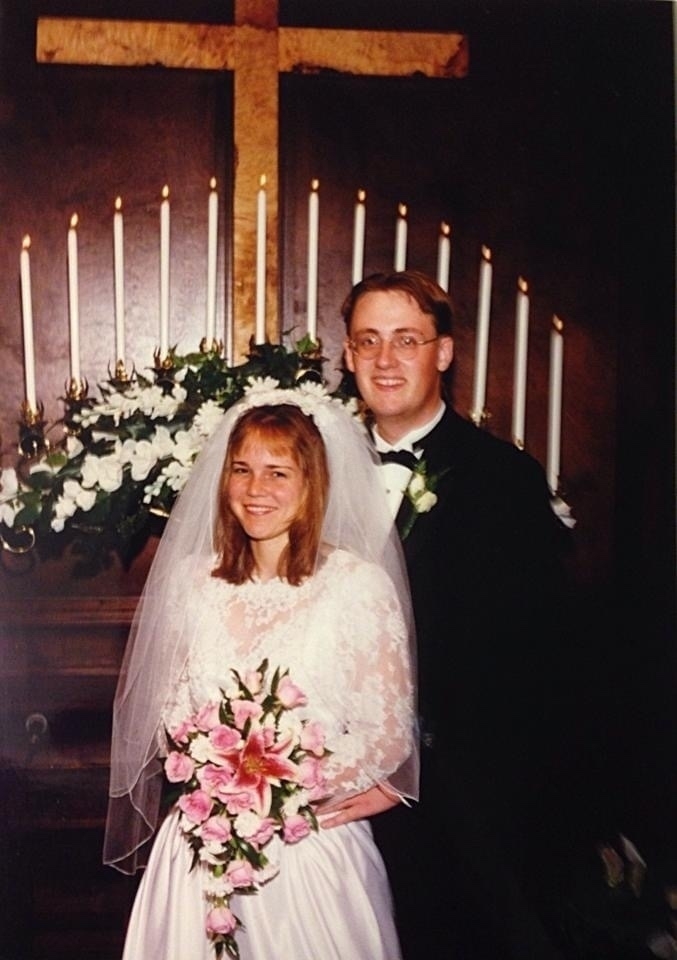 Wedding photo of a young man in a tux and a young woman in a white bridal dress standing in front of a candelabra. 