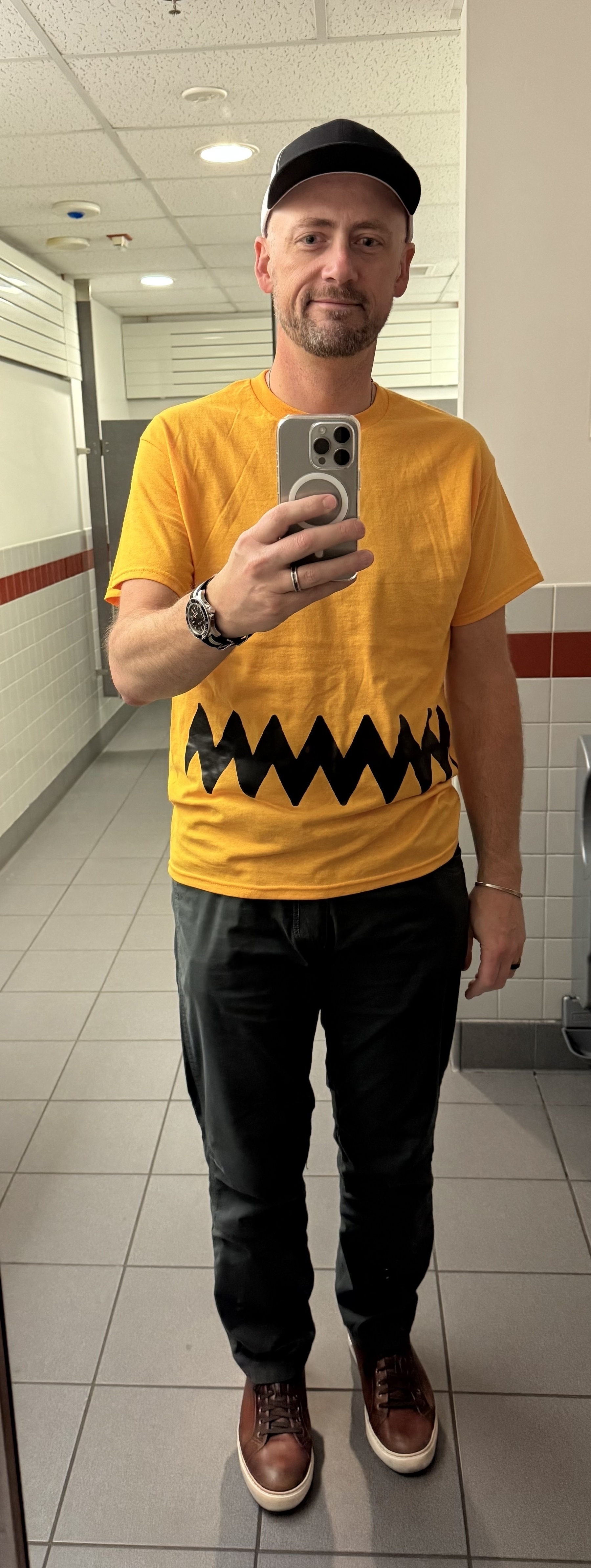 Mirror selfie of Chris in a yellow Charlie Brown T shirt with the black squiggle across the bottom, black pants, brown shoes, and a ball cap. 