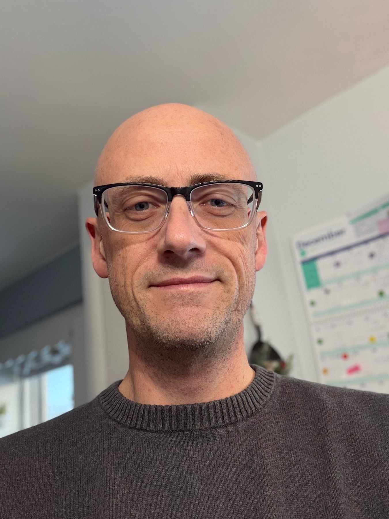 A selfie of Chris, a bald white man who is smiling and wearing a grey sweater and black-and-clear framed glasses. 