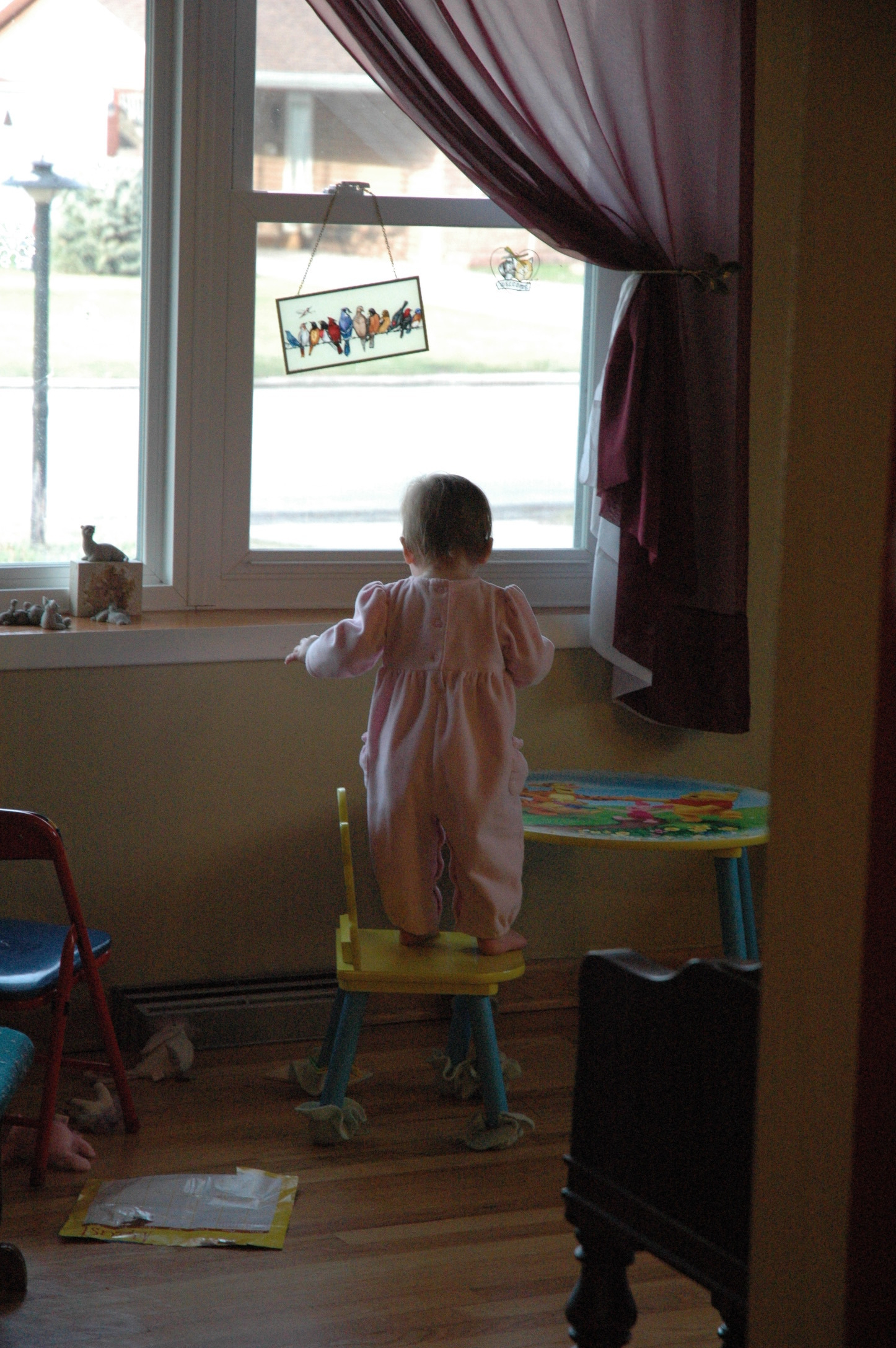 A small child wearing pajamas stands on a chair looking out the window.
