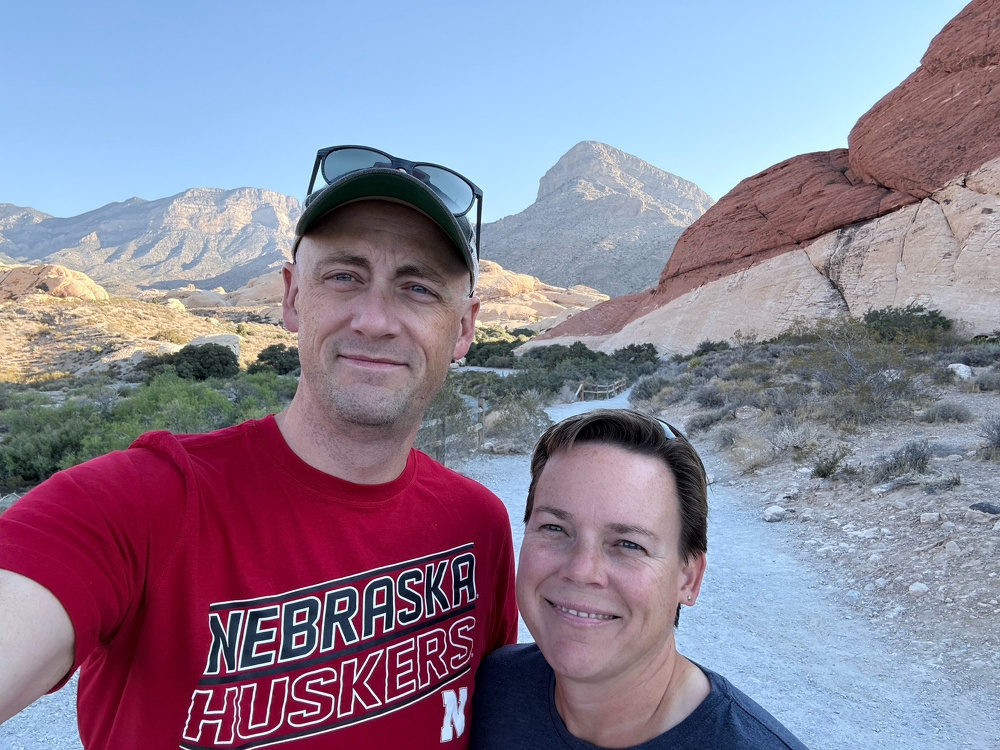 A tall man wearing a ball cap and a shorter woman with a pretty smile post for a selfie in front of a desert landscape