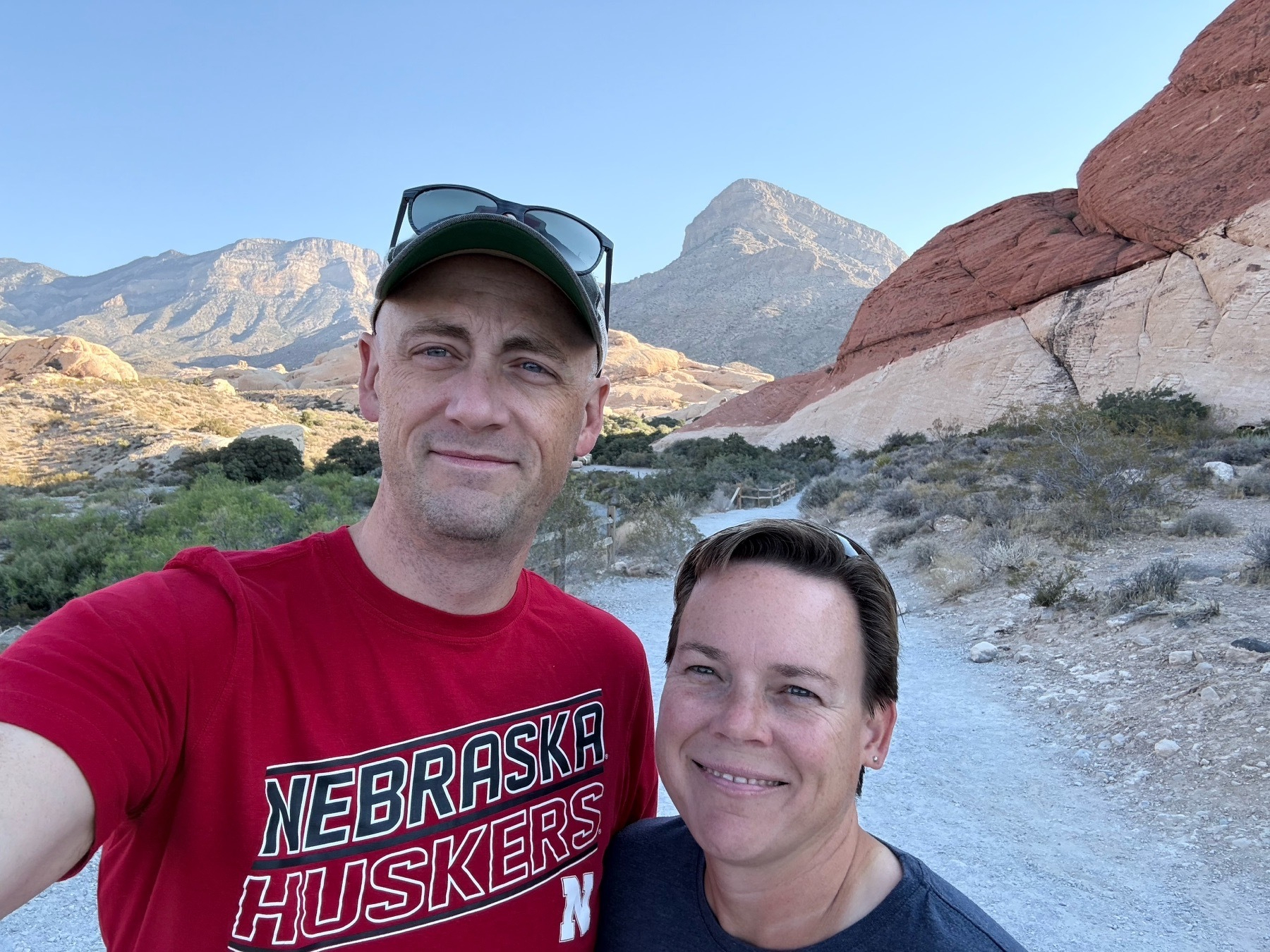 A tall man wearing a ball cap and a shorter woman with a pretty smile post for a selfie in front of a desert landscape
