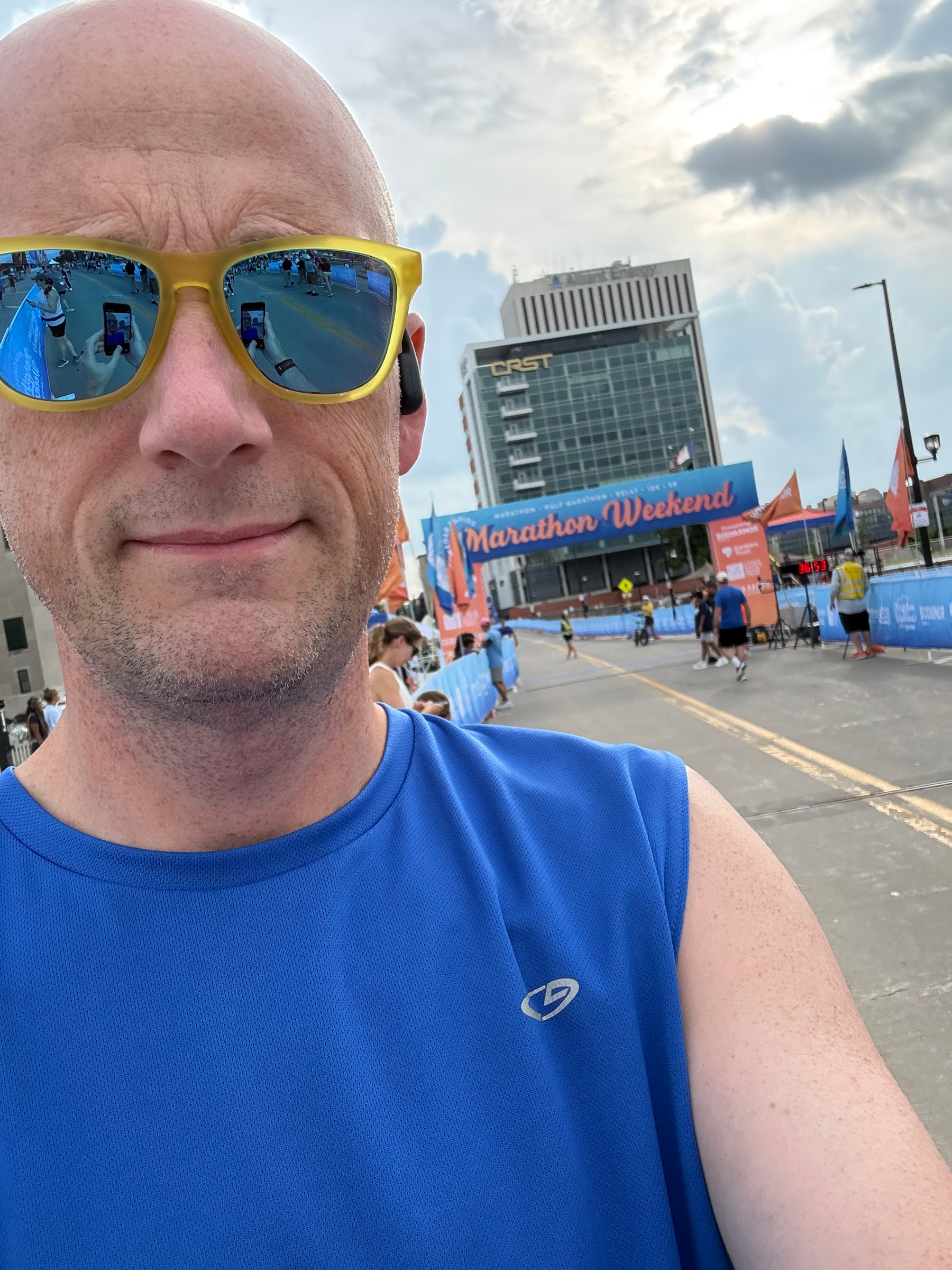 A person wearing sunglasses and a blue sleeveless shirt stands near a marathon event with other participants in the background.
