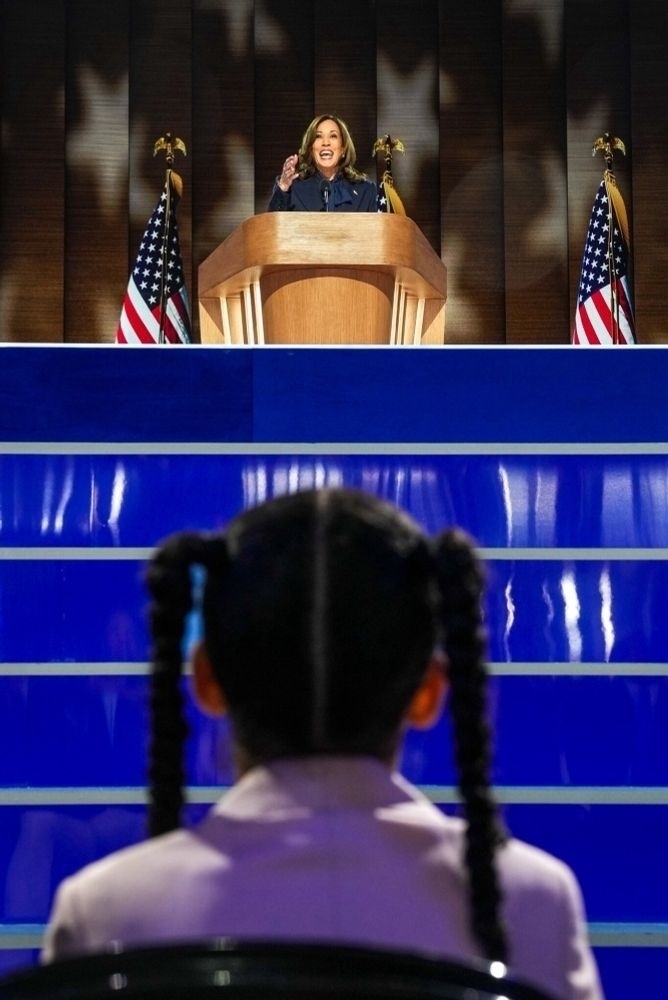 Kamala Harris on the stage at the united center. In front of and below her is the back of a young girl with pigtails in her hair.