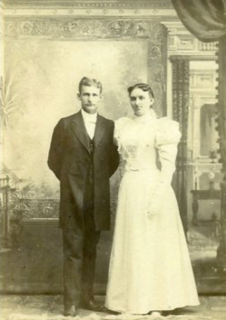 Auto-generated description: A vintage photograph features a man and a woman in formal attire, posing in front of an ornate backdrop.