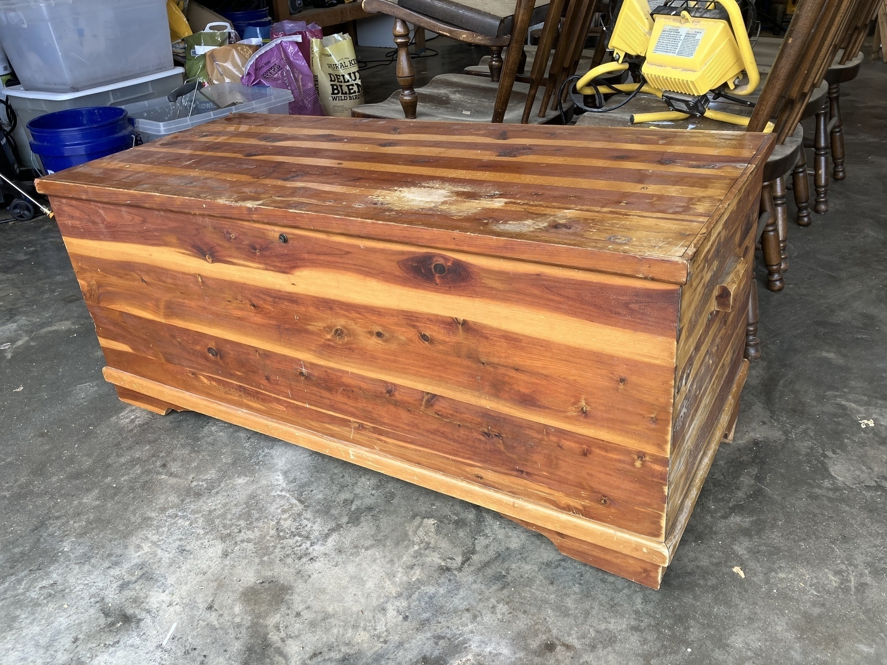 Auto-generated description: A wooden chest with a rustic appearance is placed on a concrete floor, surrounded by some chairs and a work light.