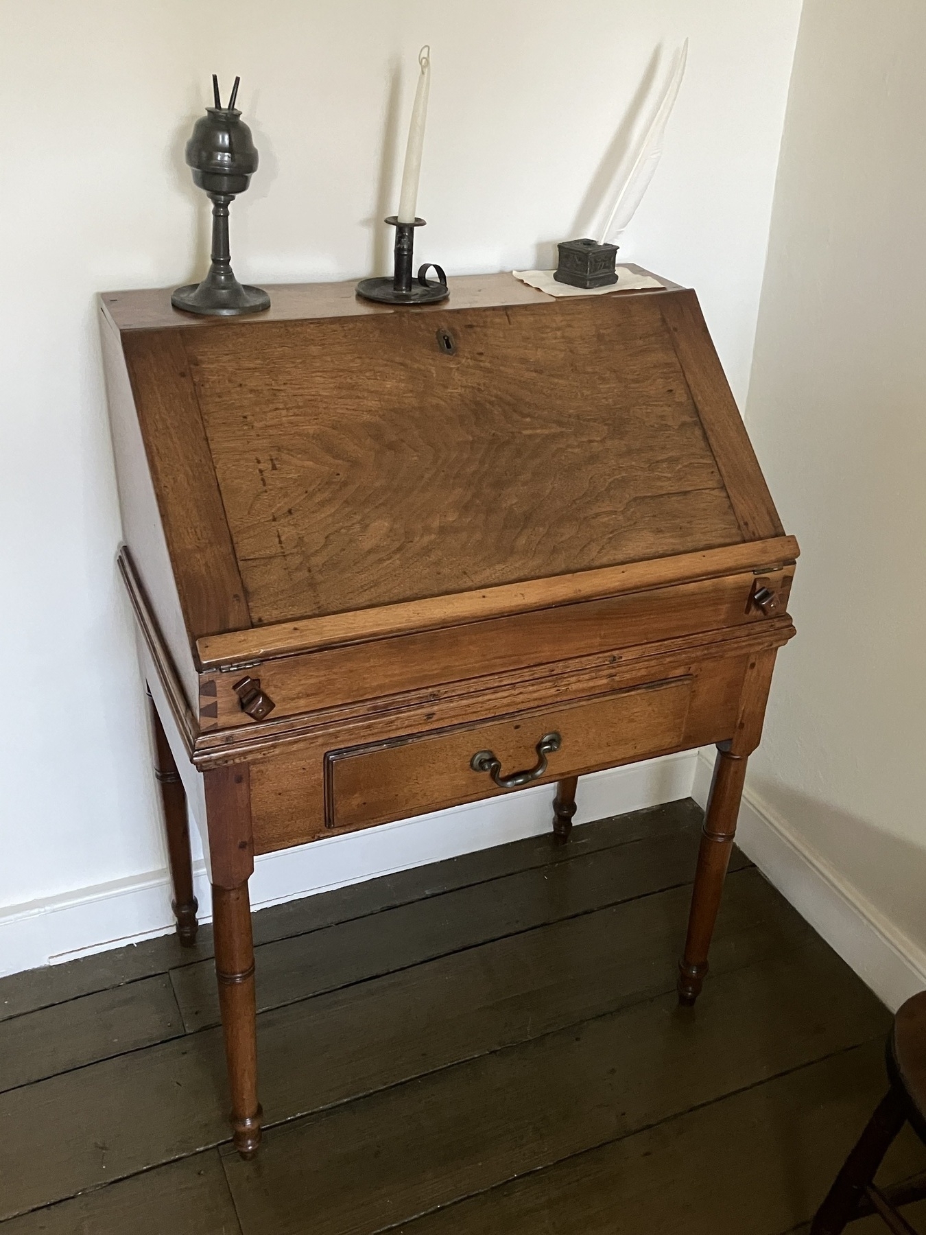 Auto-generated description: A vintage wooden writing desk with a slanted lid, a drawer below, and two candlesticks and an inkwell on top.