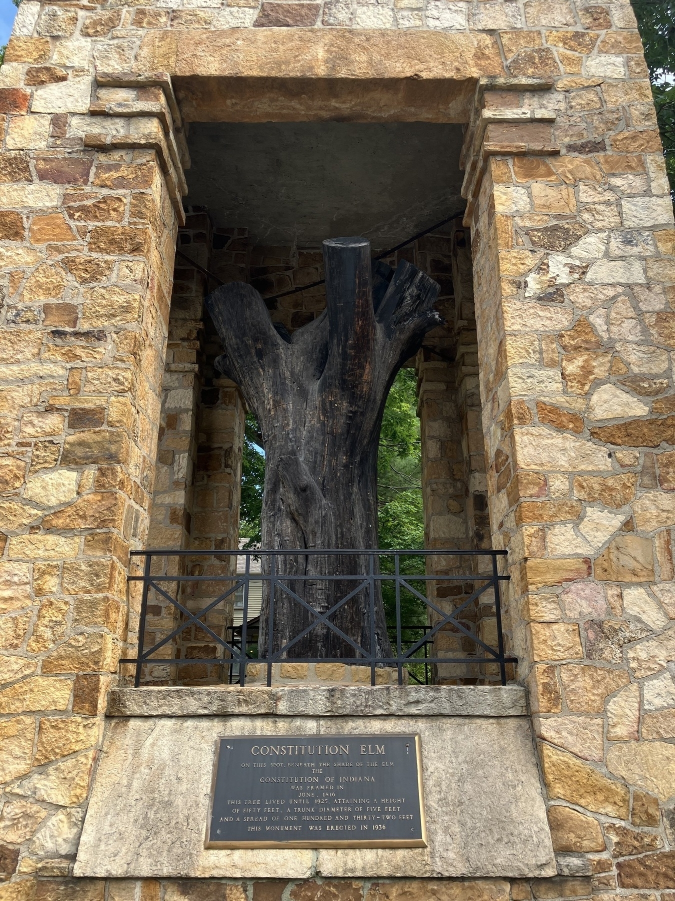 Auto-generated description: A tall wooden stump, encased in a brick and stone structure and accompanied by a commemorative plaque, stands prominently.