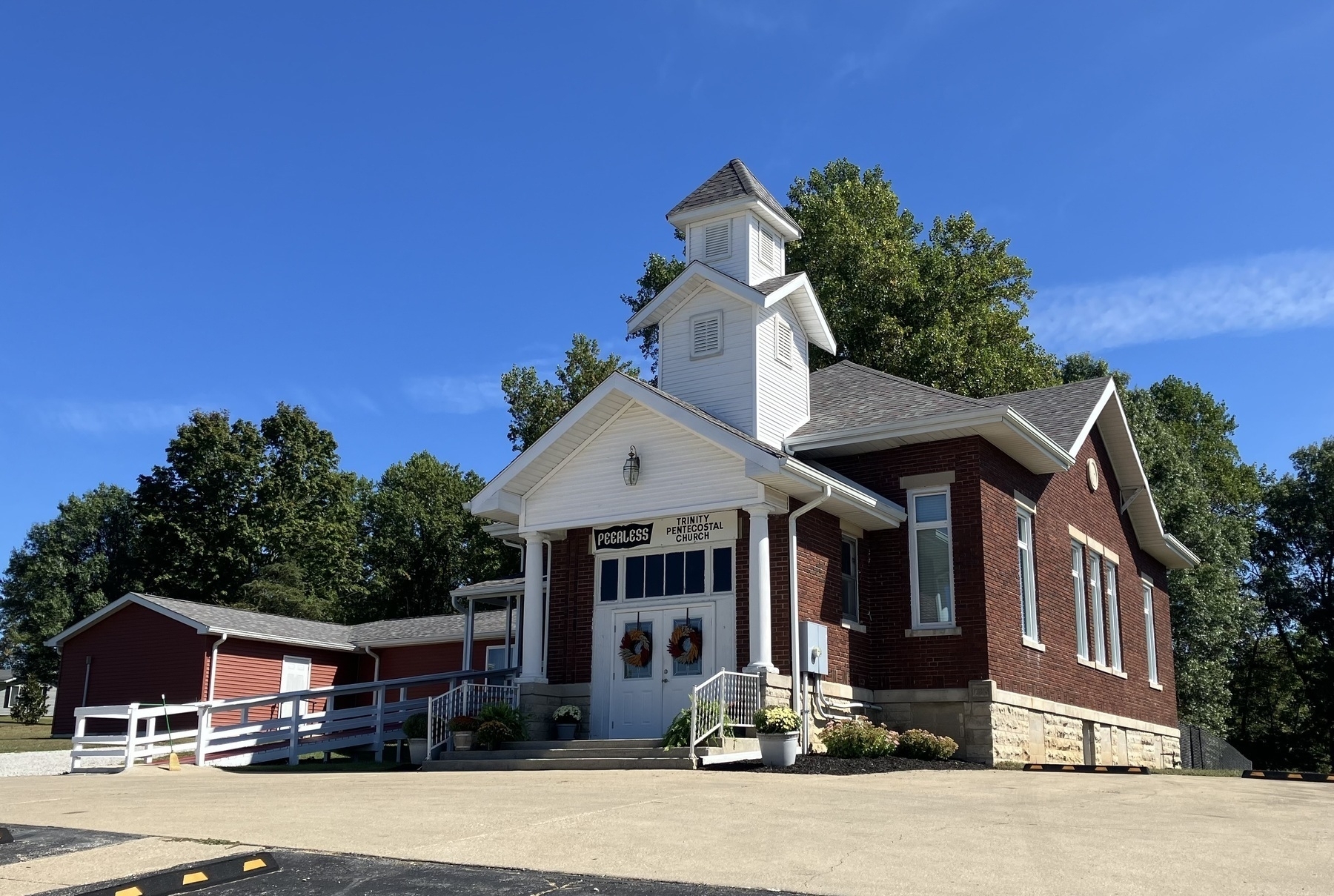 Auto-generated description: A red brick church with a white steeple is set against a clear blue sky, surrounded by greenery.