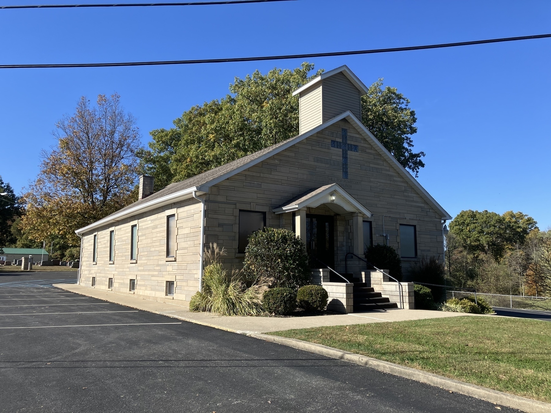 Auto-generated description: A small church building with a prominent cross, surrounded by trees, is situated next to an empty parking lot under a clear blue sky.