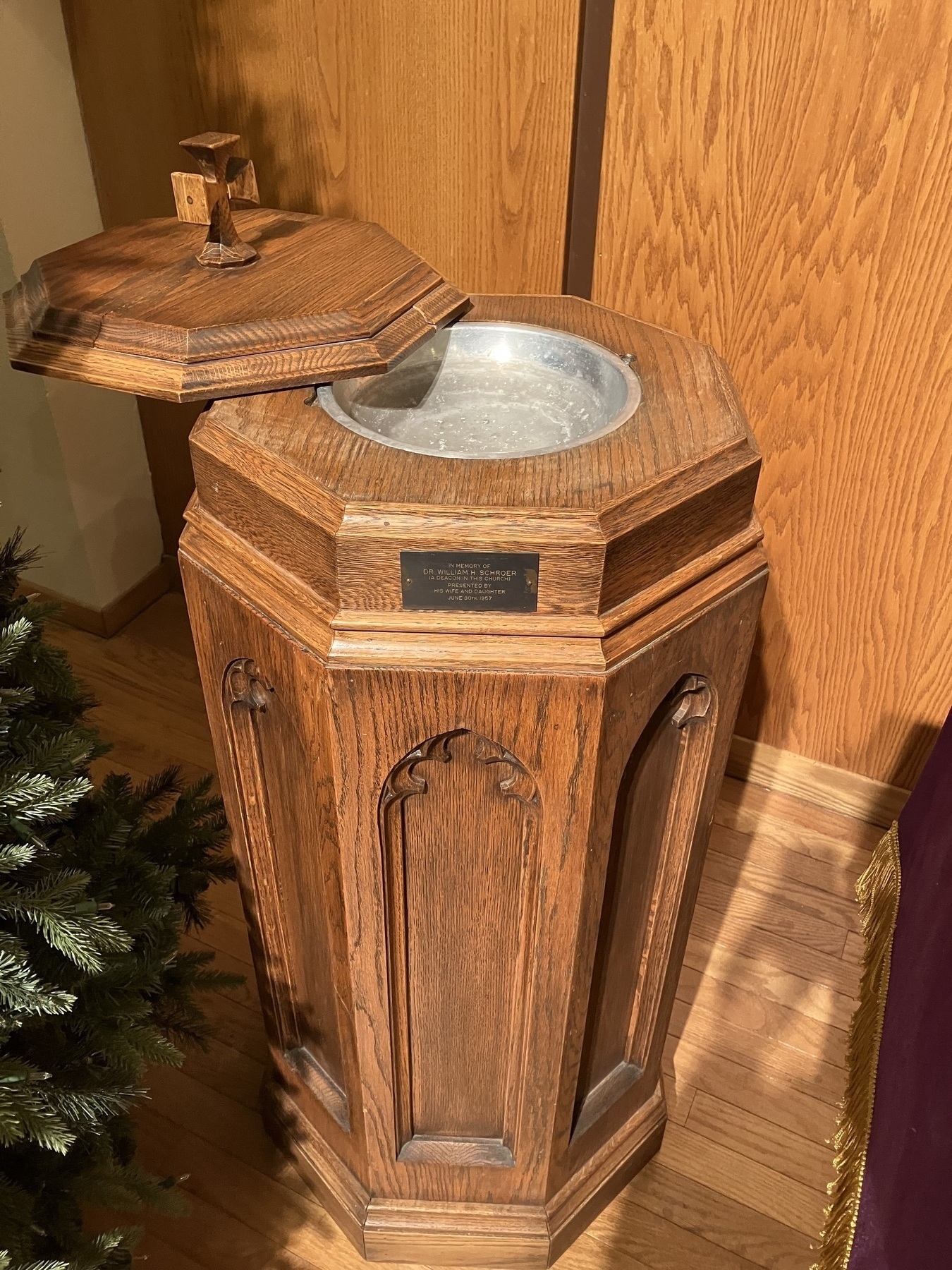 Auto-generated description: A wooden baptismal font with a hinged lid stands in a church setting, partially surrounded by greenery.