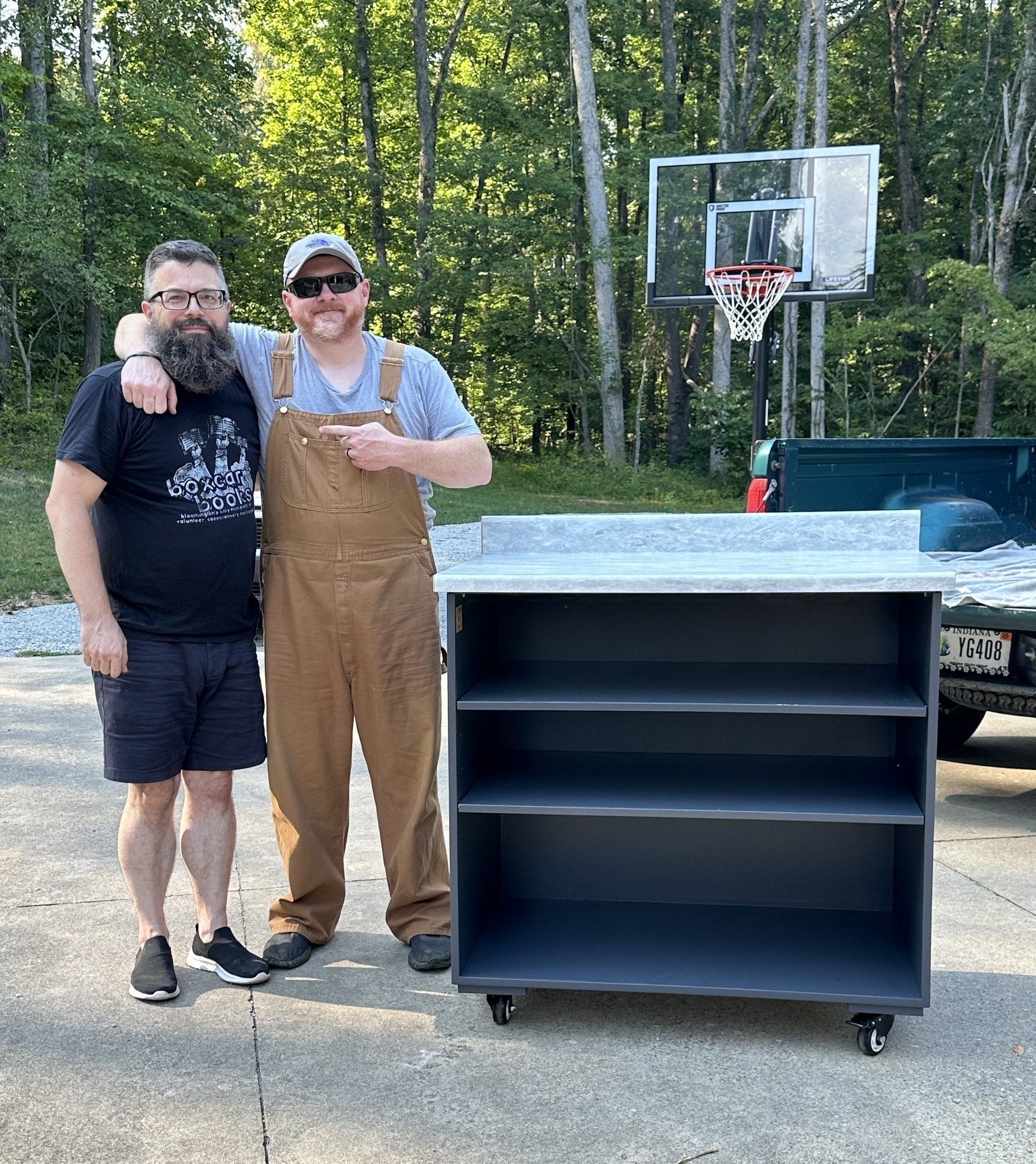 Auto-generated description: Two men are standing beside a newly-built kitchen island, smiling and posing for a photo outside near a driveway and basketball hoop.