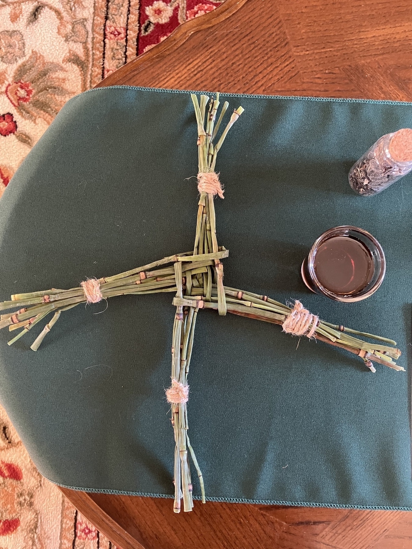 Auto-generated description: A woven cross made of straw is placed on a fabric surface alongside a small jar and a cup.