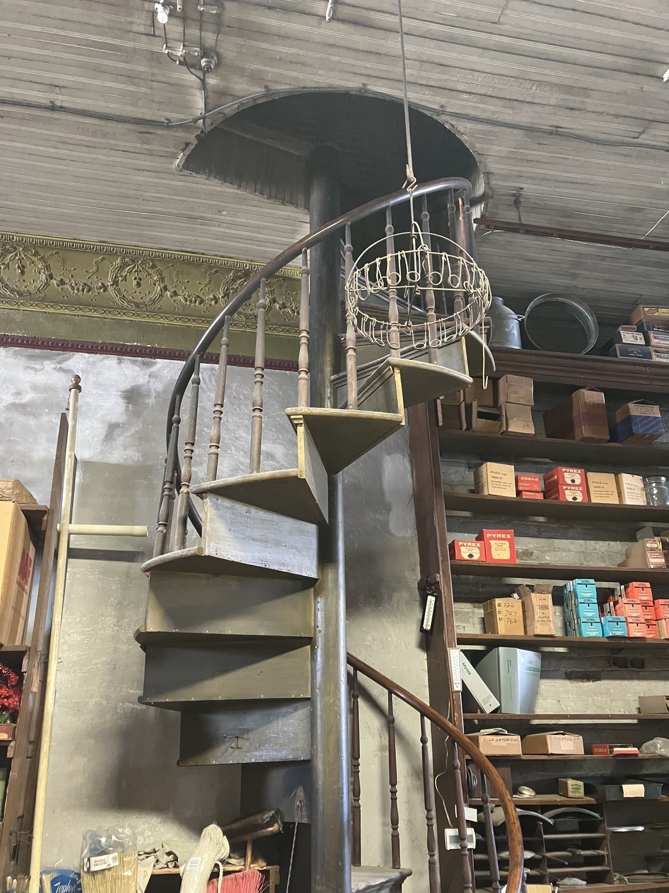 Auto-generated description: A spiral staircase with narrow steps leads up to a circular opening in the ceiling of a cluttered space filled with shelves of boxes and various items.