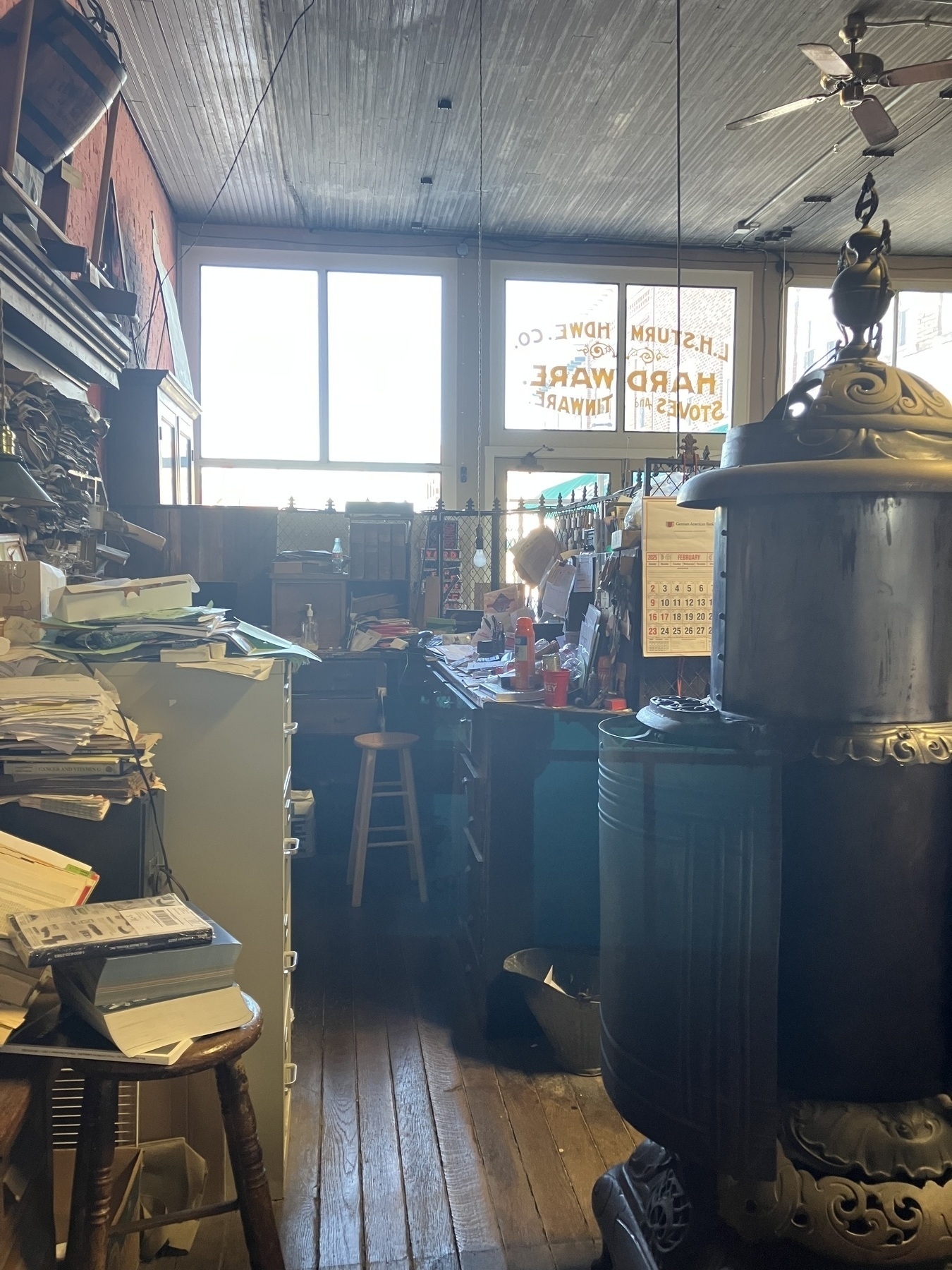 Auto-generated description: A cluttered antiques or hardware store interior is shown, featuring a large vintage stove, wooden floors, and stacks of papers, with light filtering through large windows.