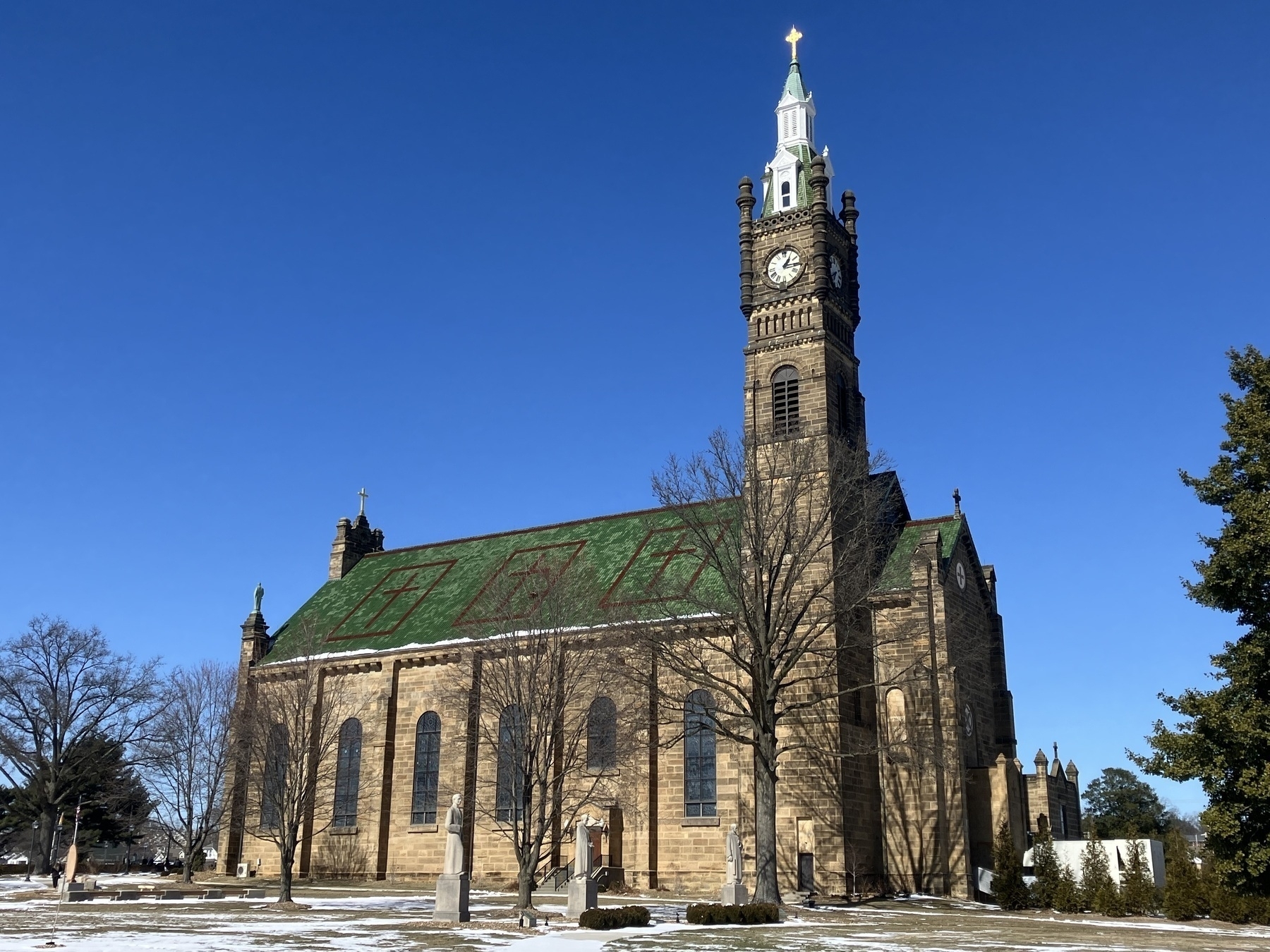 Auto-generated description: A large stone church with a clock tower and green roof stands against a clear blue sky, surrounded by some trees and snow on the ground.