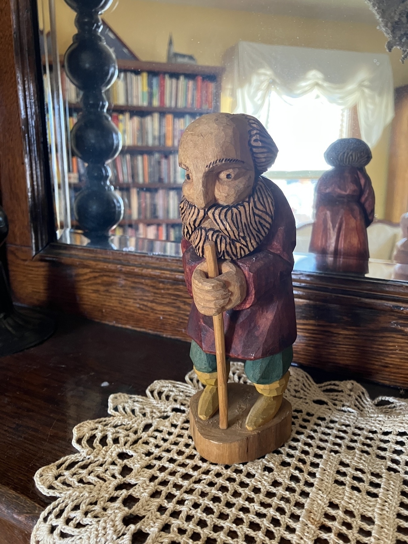 Auto-generated description: A wooden figurine of a bearded man holding a staff stands on a lace doily in front of a mirror, with shelves filled with books in the background.