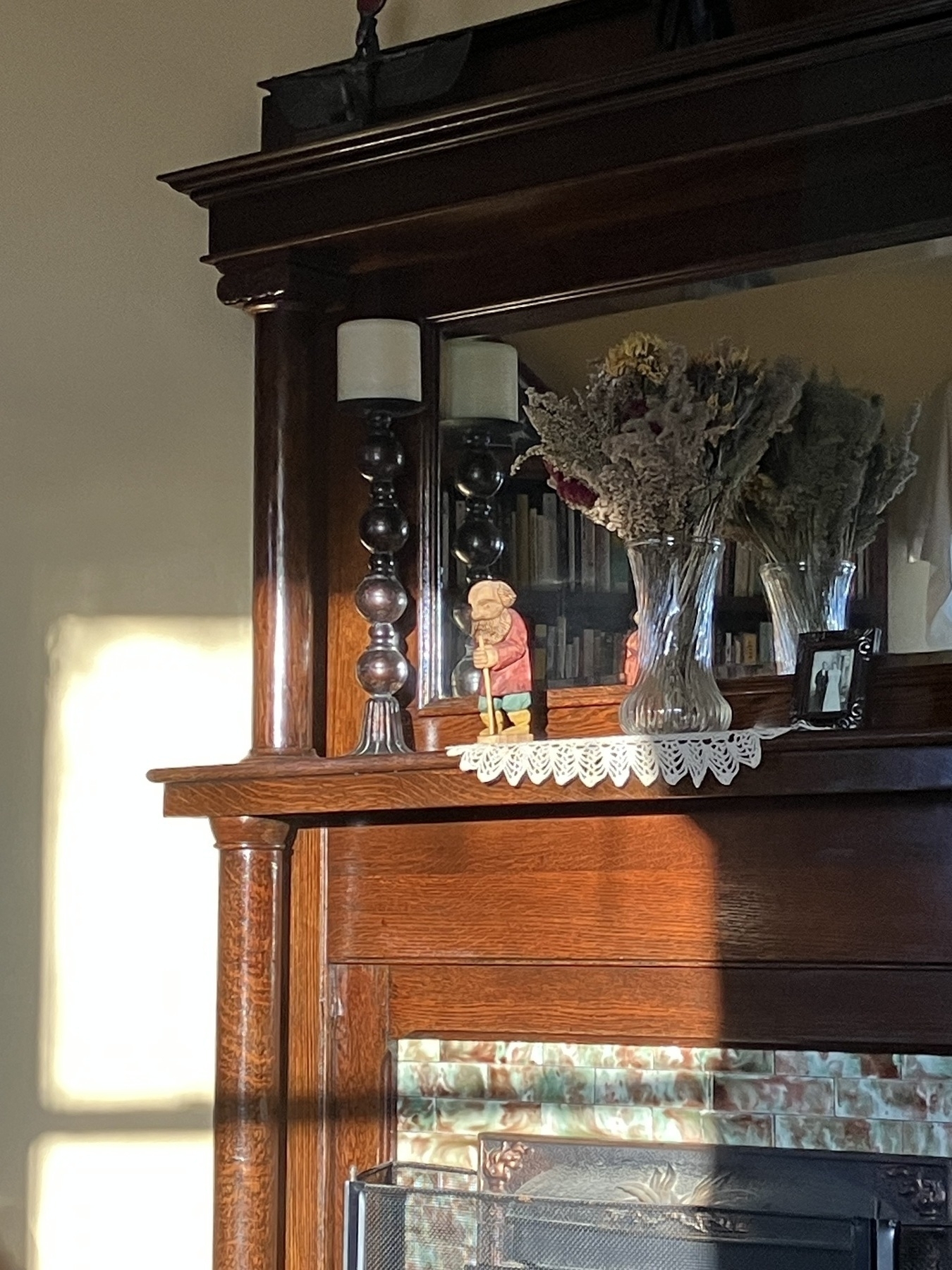 Auto-generated description: A wooden fireplace mantel is decorated with a tall candle holder, a small figure, dried flowers in a vase, and a framed photograph.