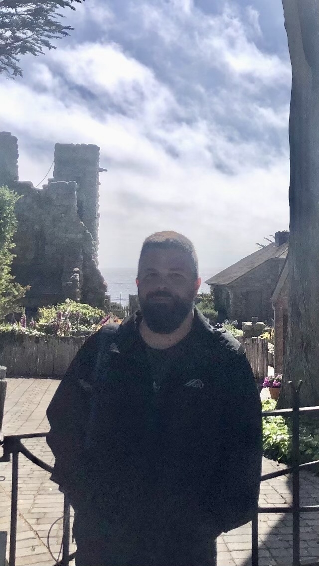 Auto-generated description: A bearded individual is standing in a sunlit outdoor setting with stone ruins and greenery in the background.