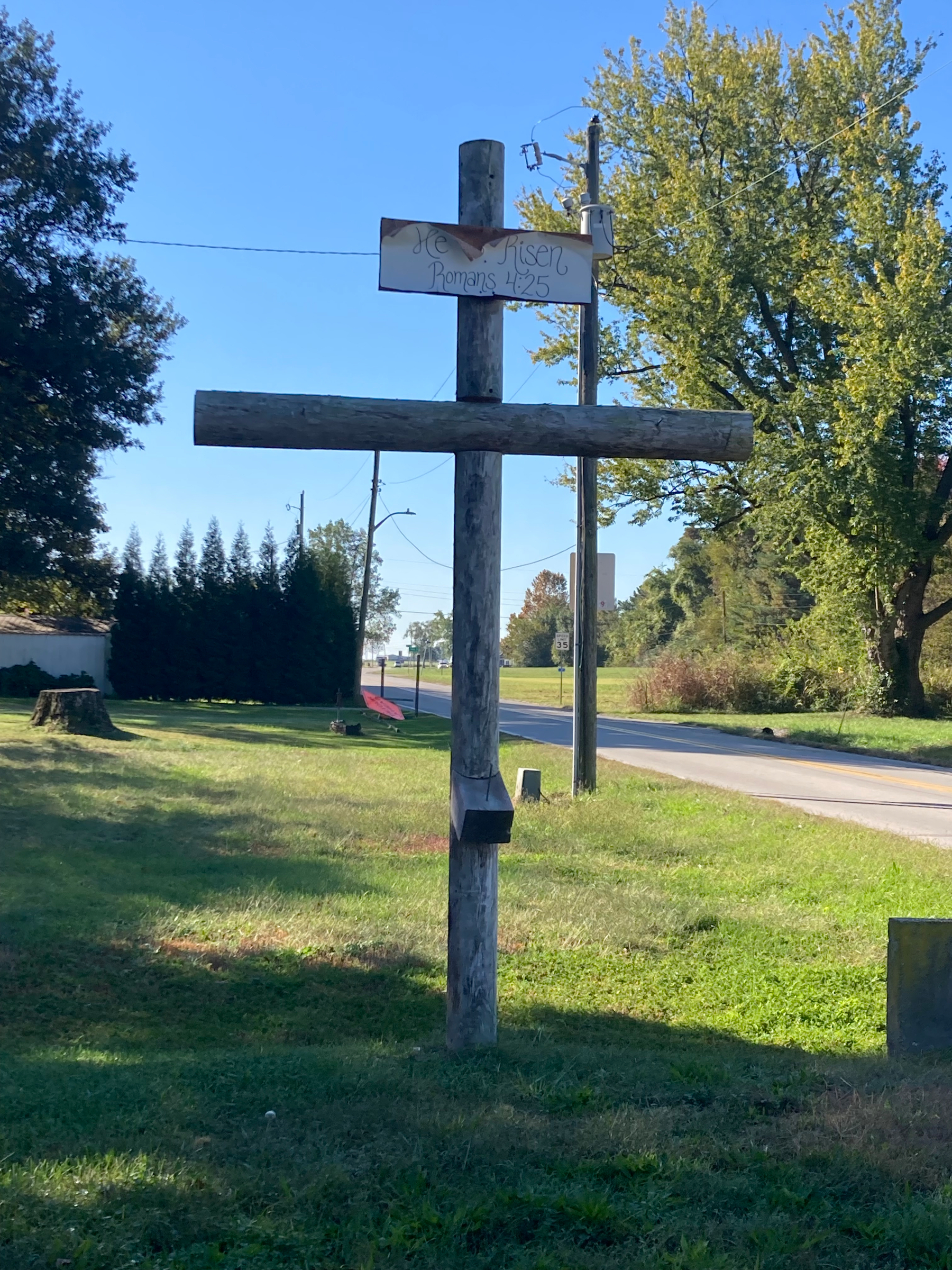 Auto-generated description: A large wooden cross stands on a grassy area near a road, with a small sign that reads He is Risen Romans 4:25 attached to it.