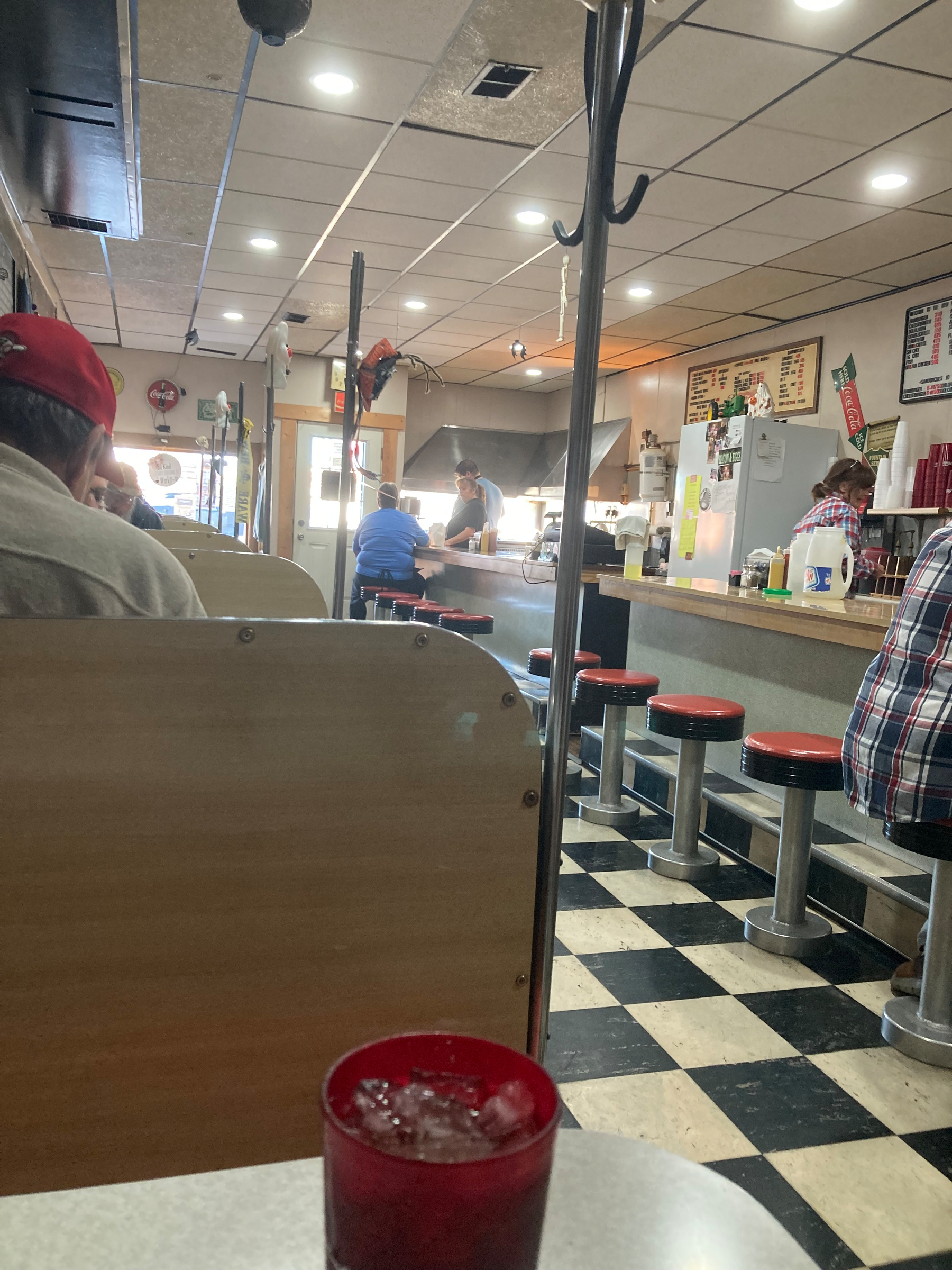 Auto-generated description: A classic diner setting with customers sitting at booths and stools along a counter, featuring a checkered floor and a red cup filled with ice on a table.