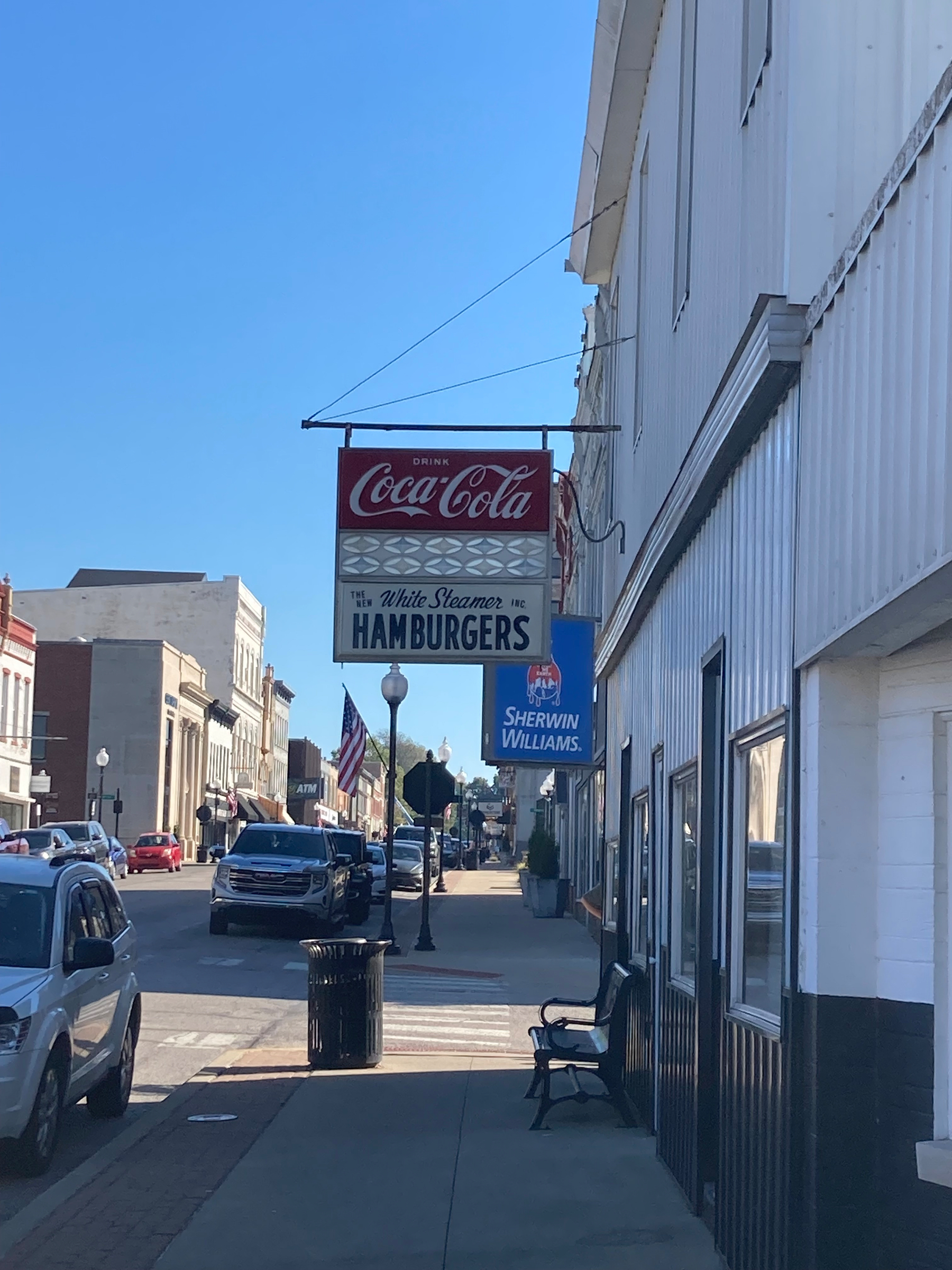 Auto-generated description: A street view shows vintage Coca-Cola and White Steamer Hamburgers signs hanging outside a building with parked cars and other shops along the sunny sidewalk.