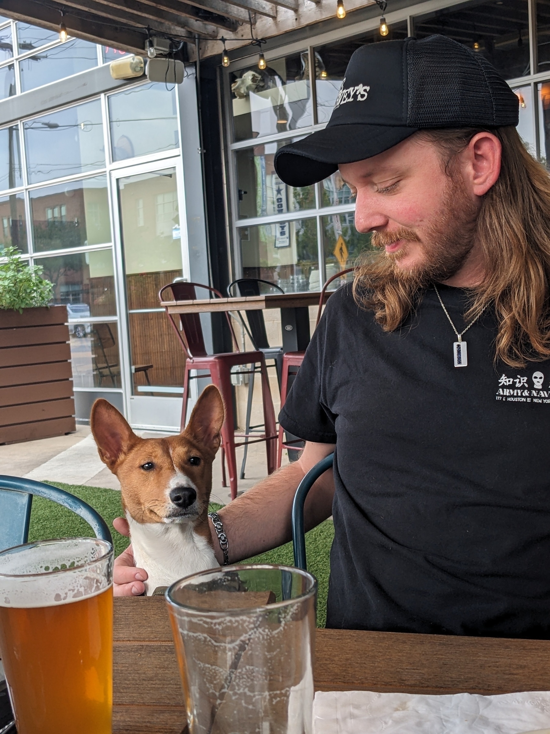 Man with long hair and beard wearing a black baseball cap and holding his Basenji dog