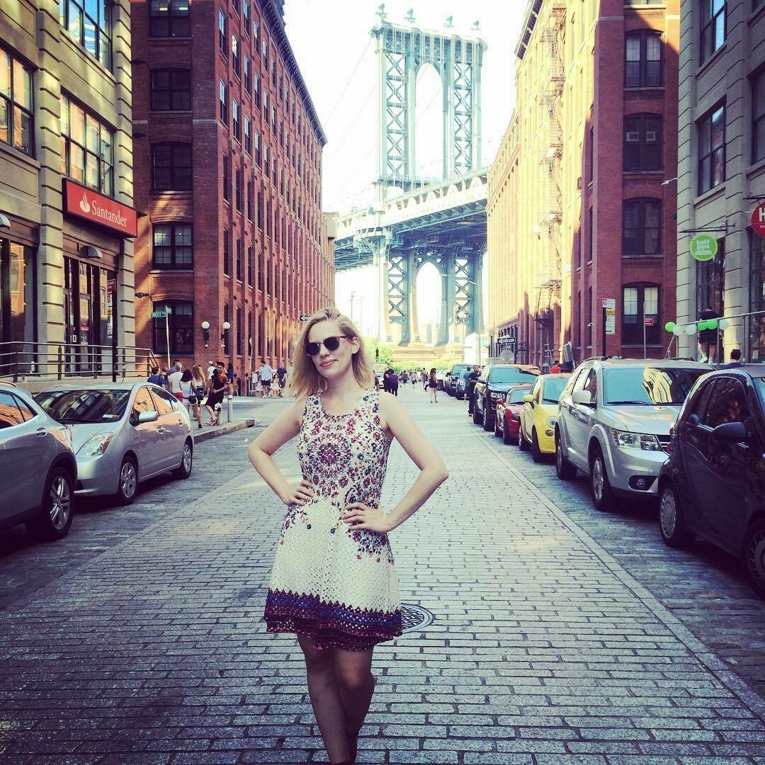 Woman in a white and patterned dress and sunglasses standing in a Brooklyn street with the Brooklyn Bridge in the background.