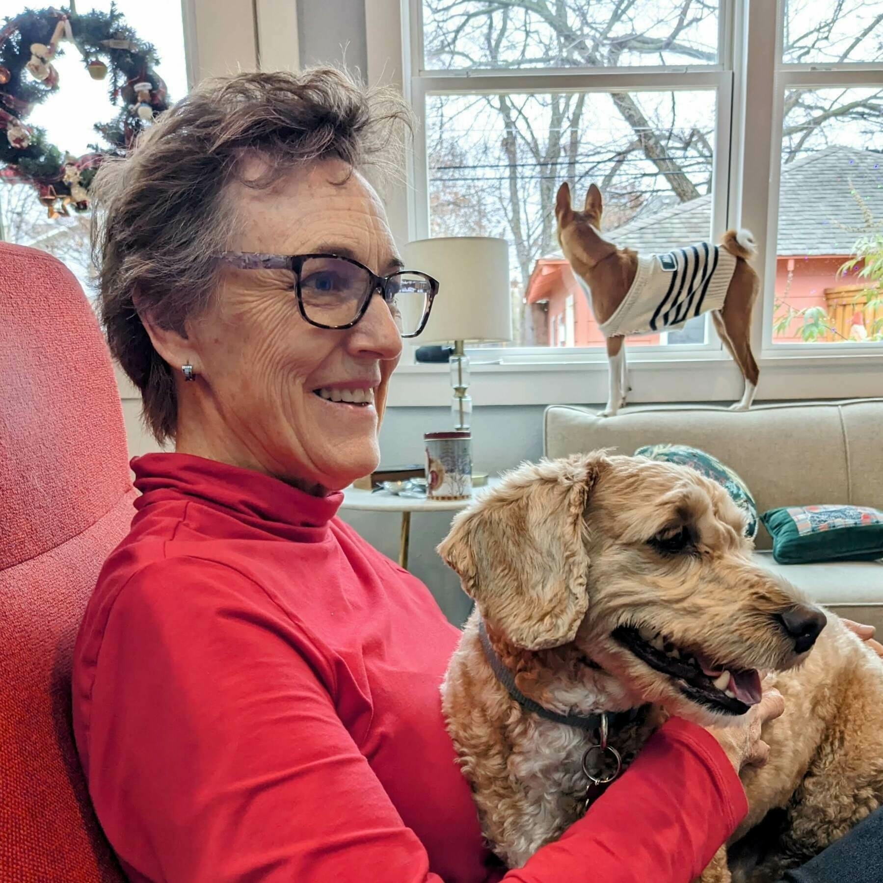 Auto-generated description: A person with glasses sits on a red chair, smiling and holding a fluffy dog, with another dog wearing a sweater standing on the couch in the background.