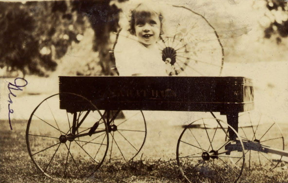 Auto-generated description: A young girl is sitting in a vintage wagon holding a parasol.