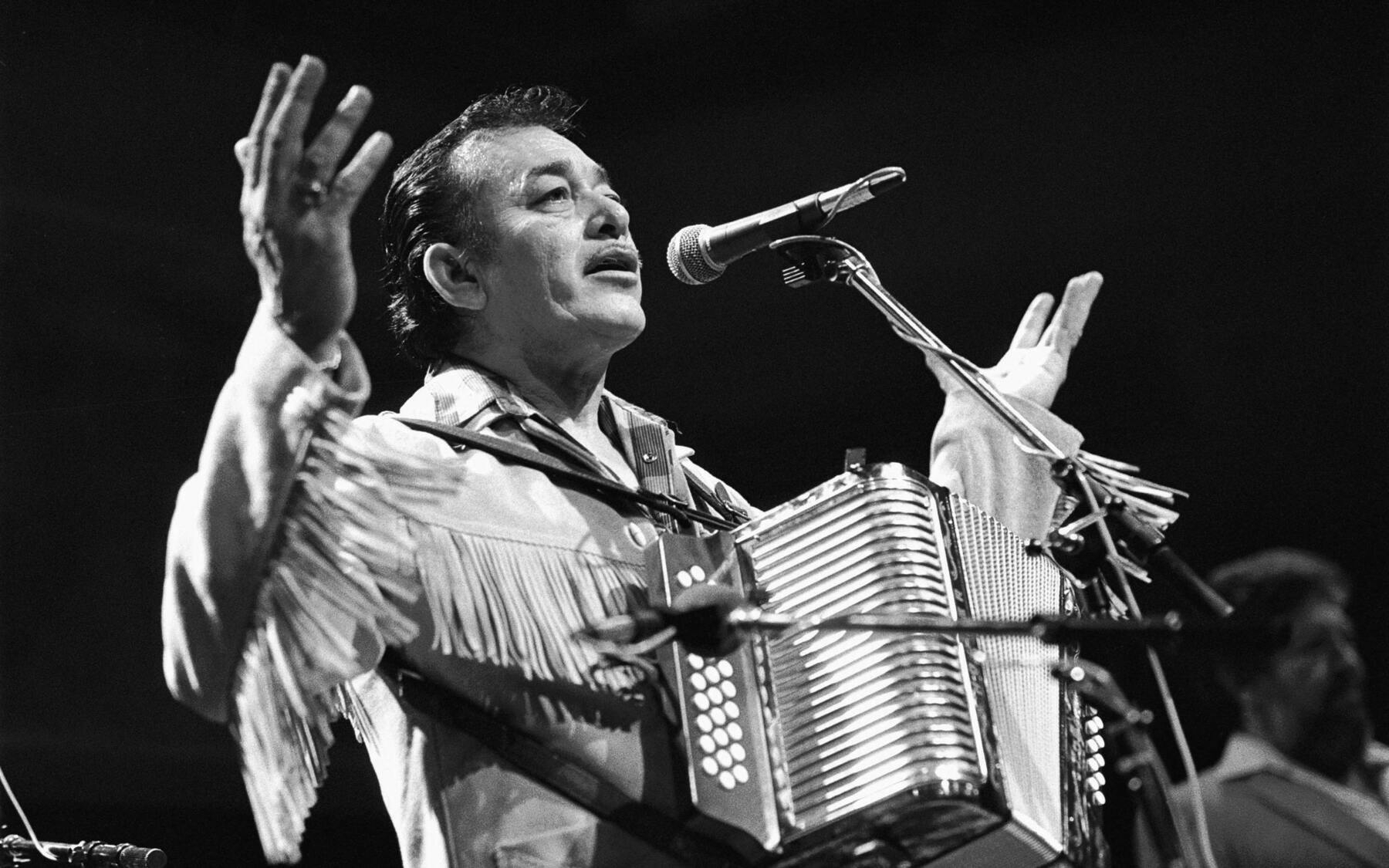 The great Flaco Jiménez performs with an accordion while singing into a microphone on stage.