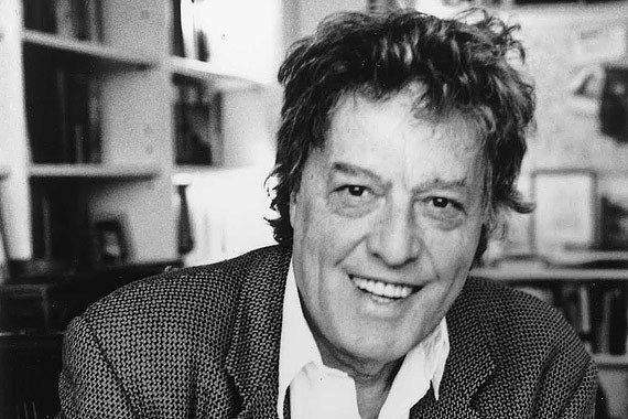 Tom Stoppard, with a joyful smile is sitting in a room with bookshelves in the background.
