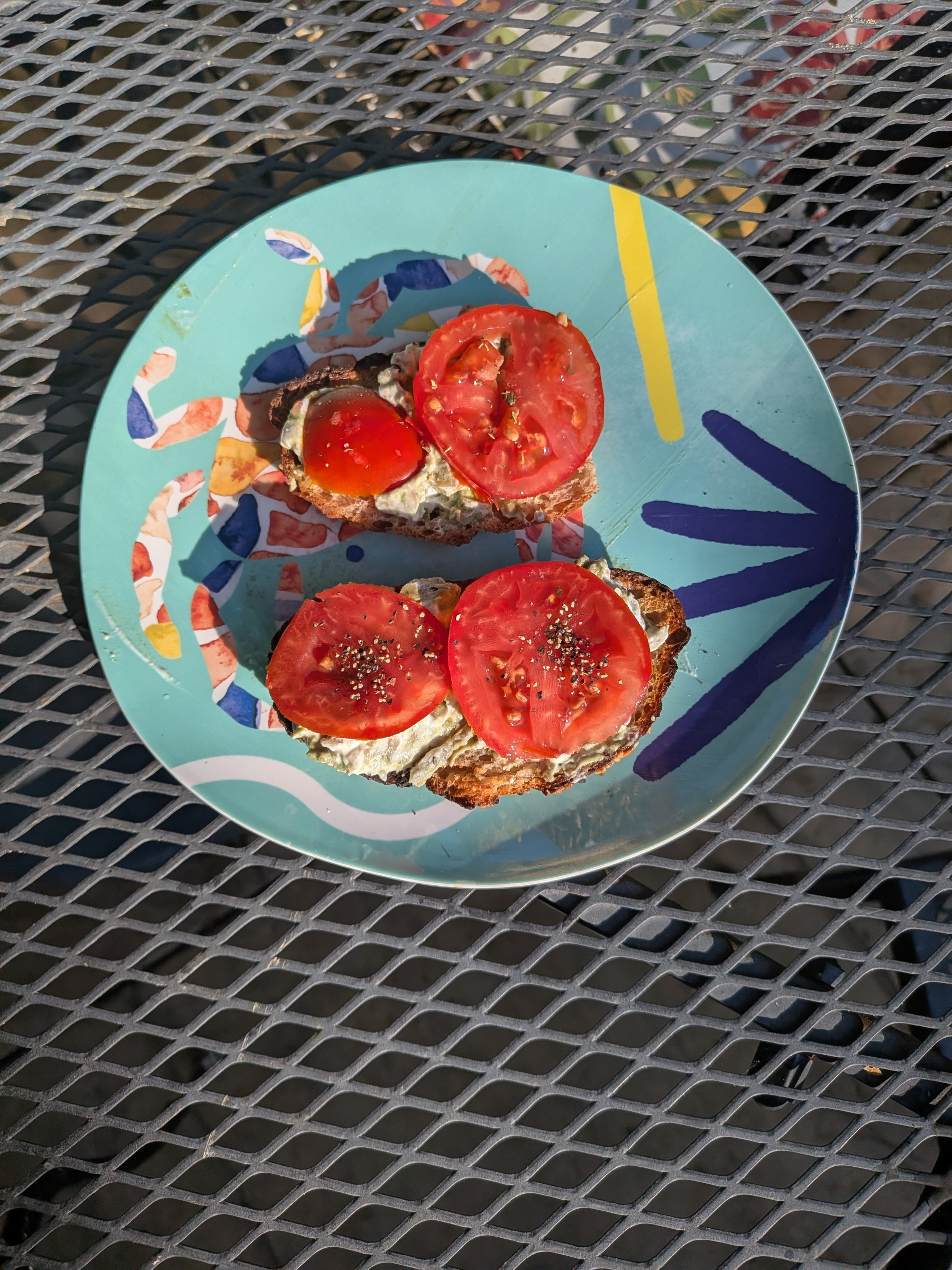 Two slices of toast with cream cheese, tomato slices, and chia seeds are placed on a colorful plate set on a metal mesh table.