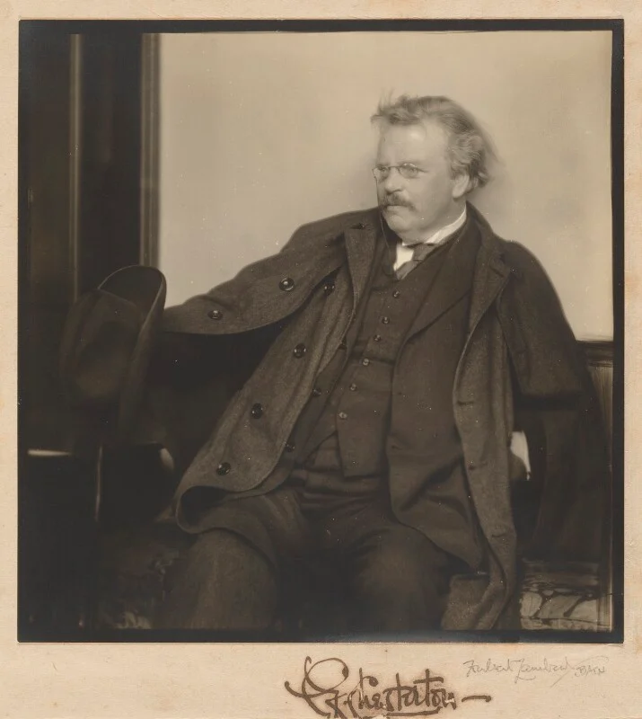The English writer, G.K. Chesterton, seated in formal attire, draped in a heavy coat, holds a hat in one hand and gazes thoughtfully to the side.