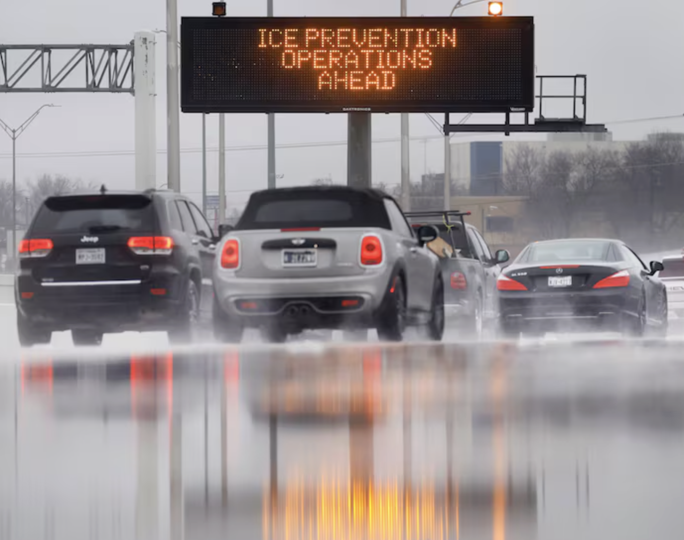 Auto-generated description: Cars drive on a wet road beneath a sign warning of ICE PREVENTION OPERATIONS AHEAD.