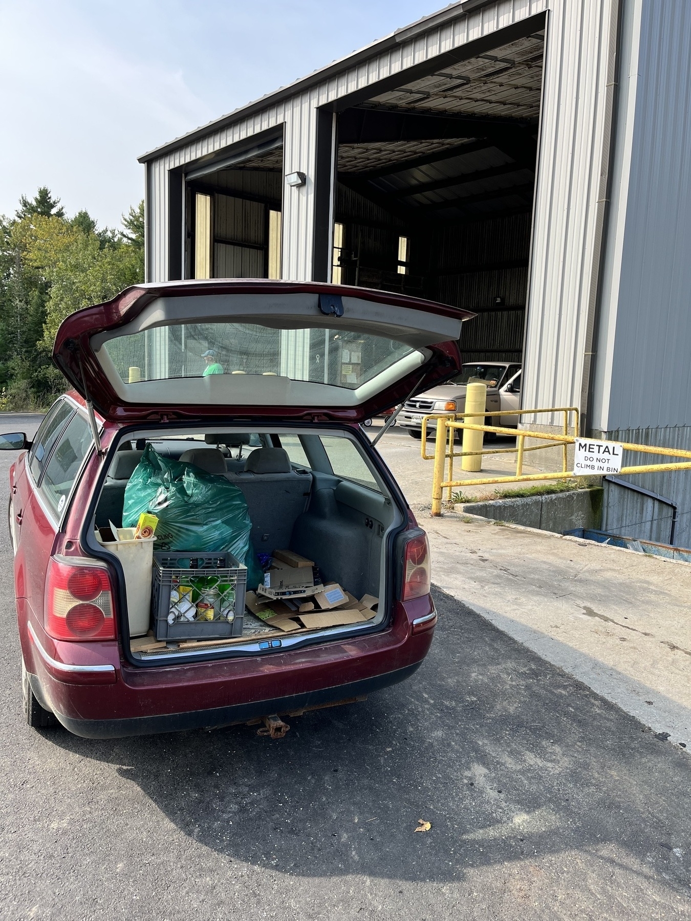 View of open back of station wagon filled with trash and recycling in front of large metal building. 