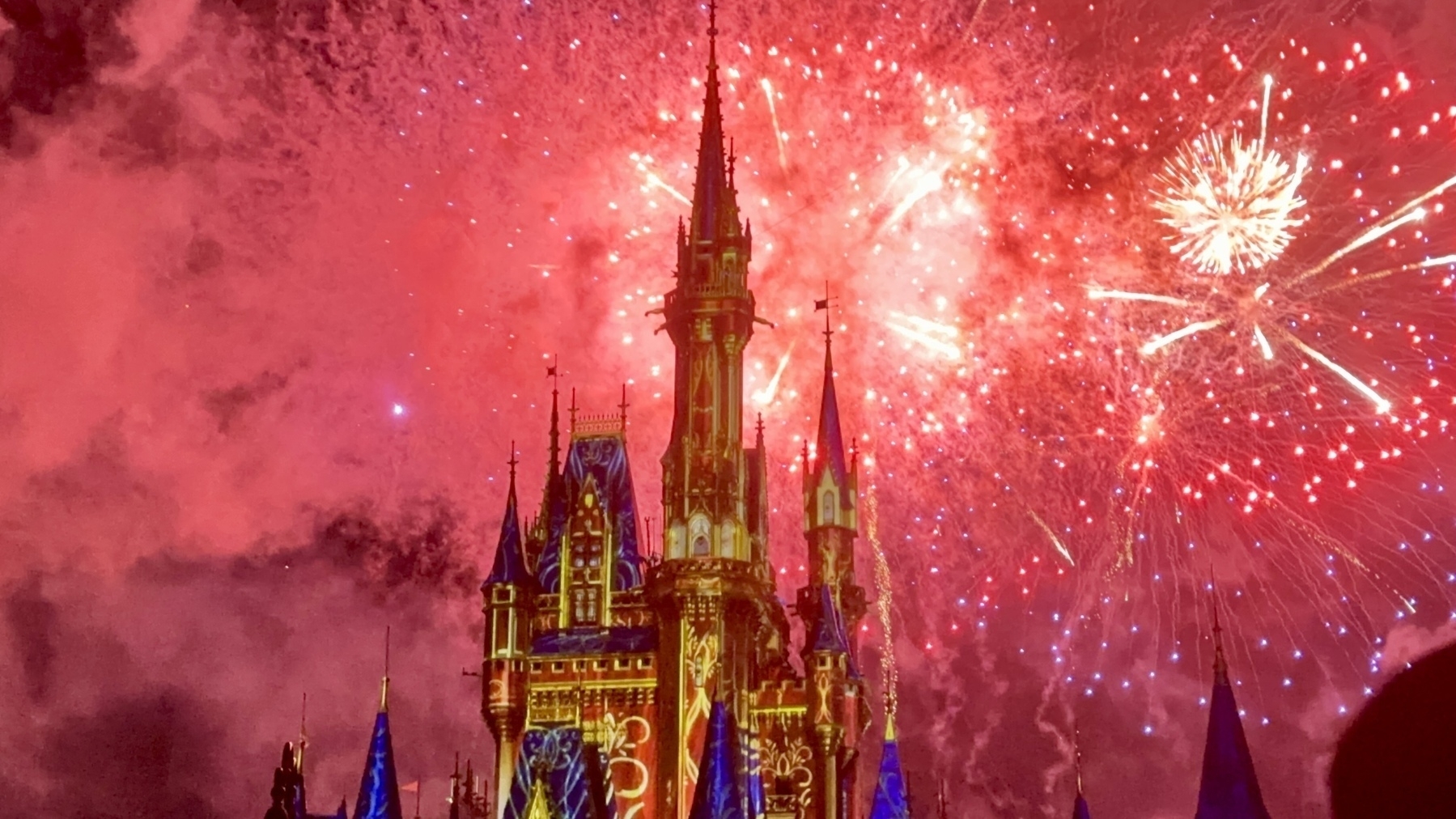 A brightly lit castle is illuminated by colorful fireworks exploding in a vibrant night sky.