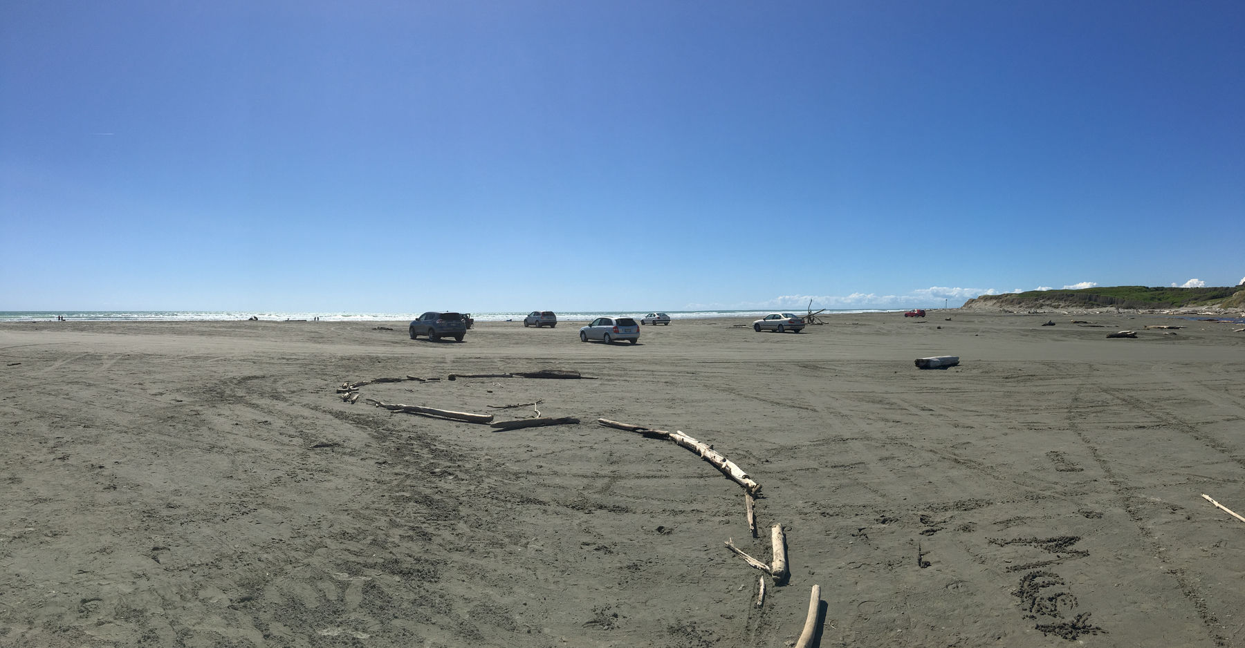 A photo of several cars parked up on a beach, with the waves crashing in the background in the mid afternoon on a beautiful late spring day