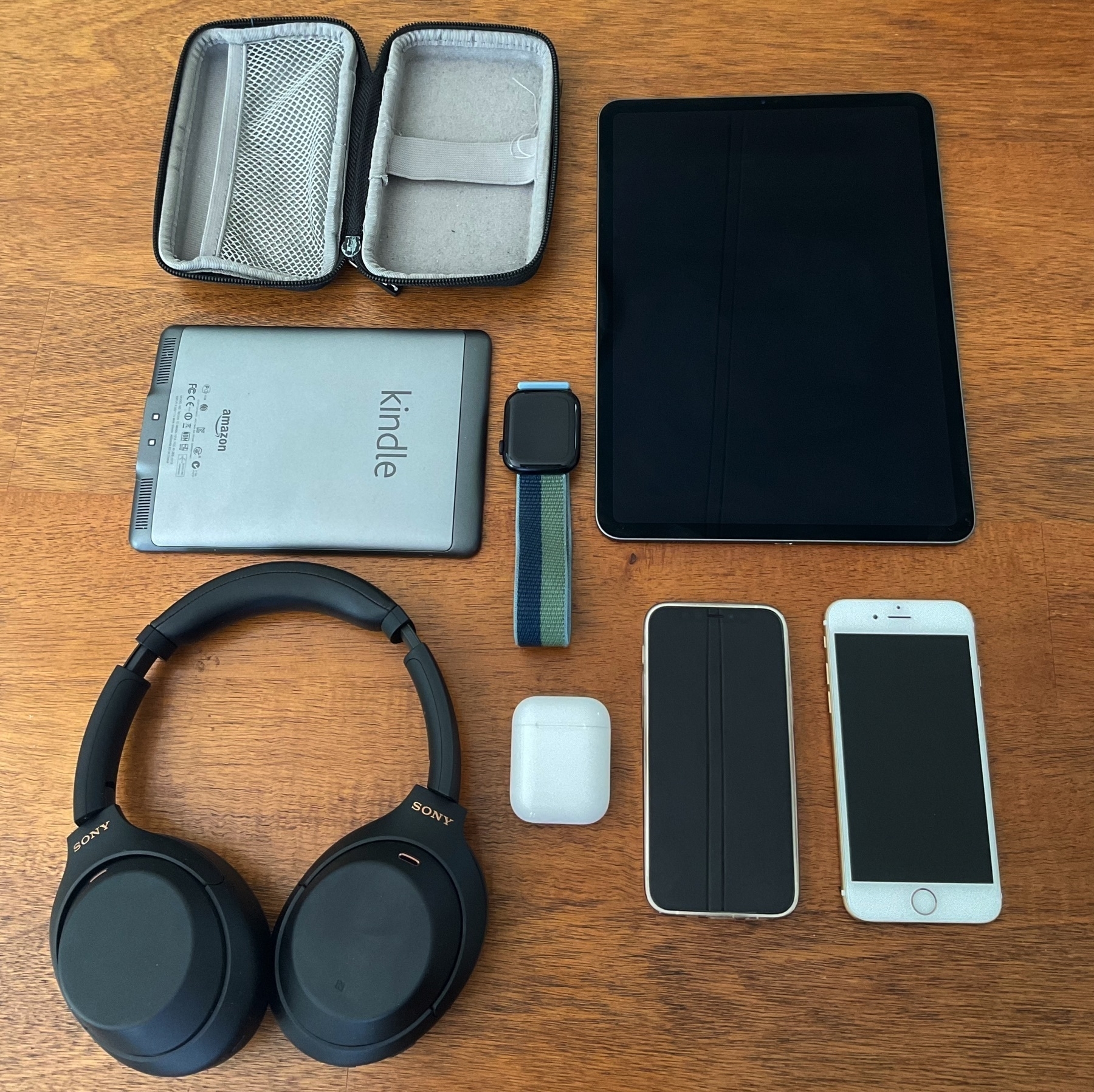 A layout of the gadgets we normally take on trips, laid out in a grid on a table: An iPad, 2 iPhones, a pair of Airpods, a pair of headphones, a kindle and an Apple Watch.