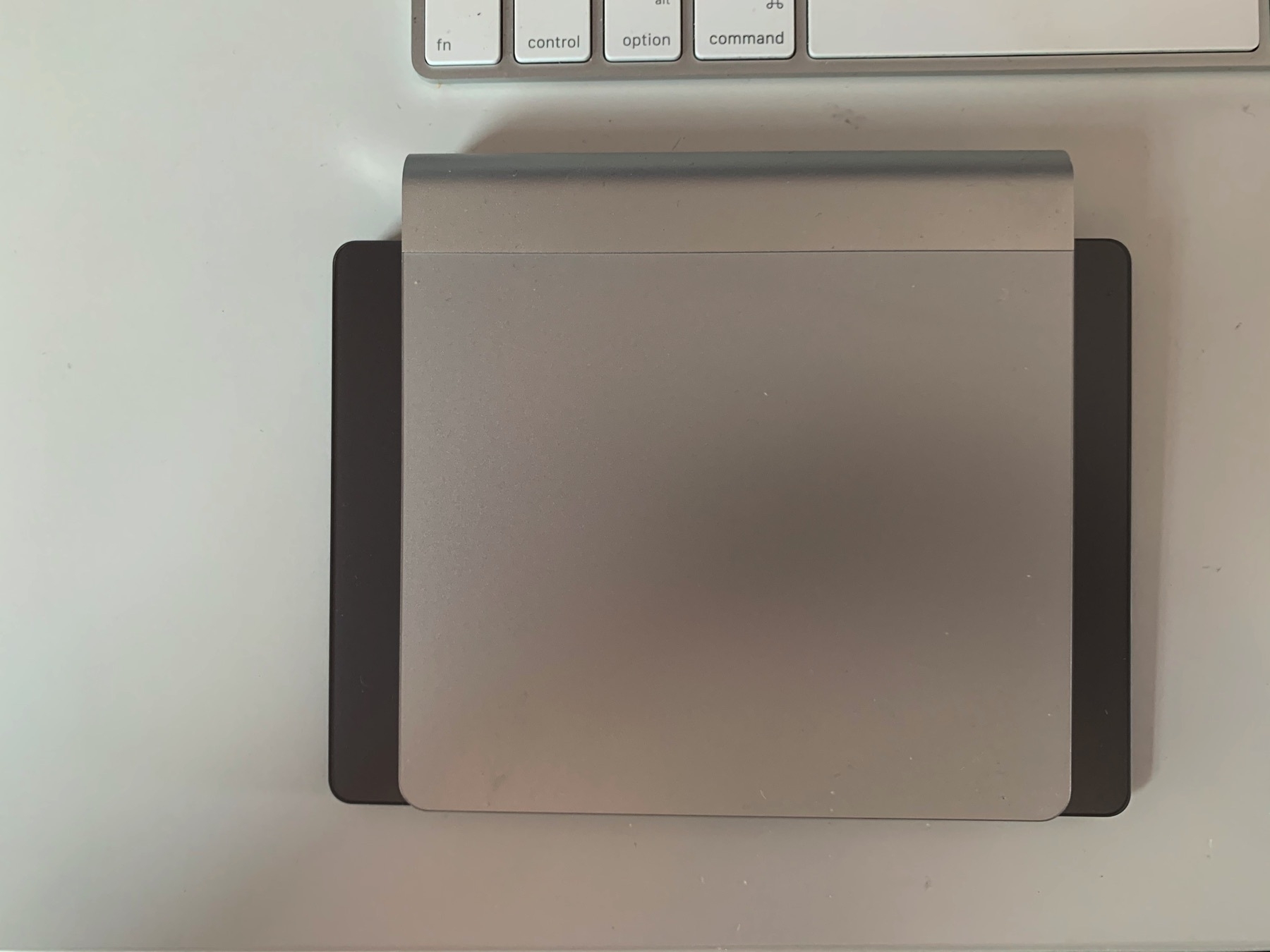 A silver original generation Magic Trackpad sitting on top of a space grey magic trackpad 2. The Magic Trackpad 2 has a much bigger surface area, and so can be seen poking out the sides below the original trackpad.
