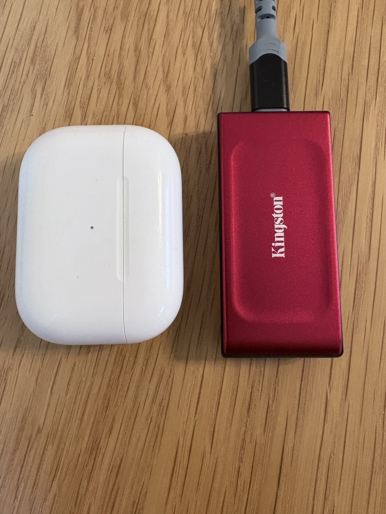 A small, bright red Kingston SSD sitting next to an AirPods Pro 2 case for size comparison. The drive is slightly longer than the case (maybe 20%?), but is much narrower (~75%) and not as tall (~80%). The drive is rectangular and has sharp, squared off edges. 